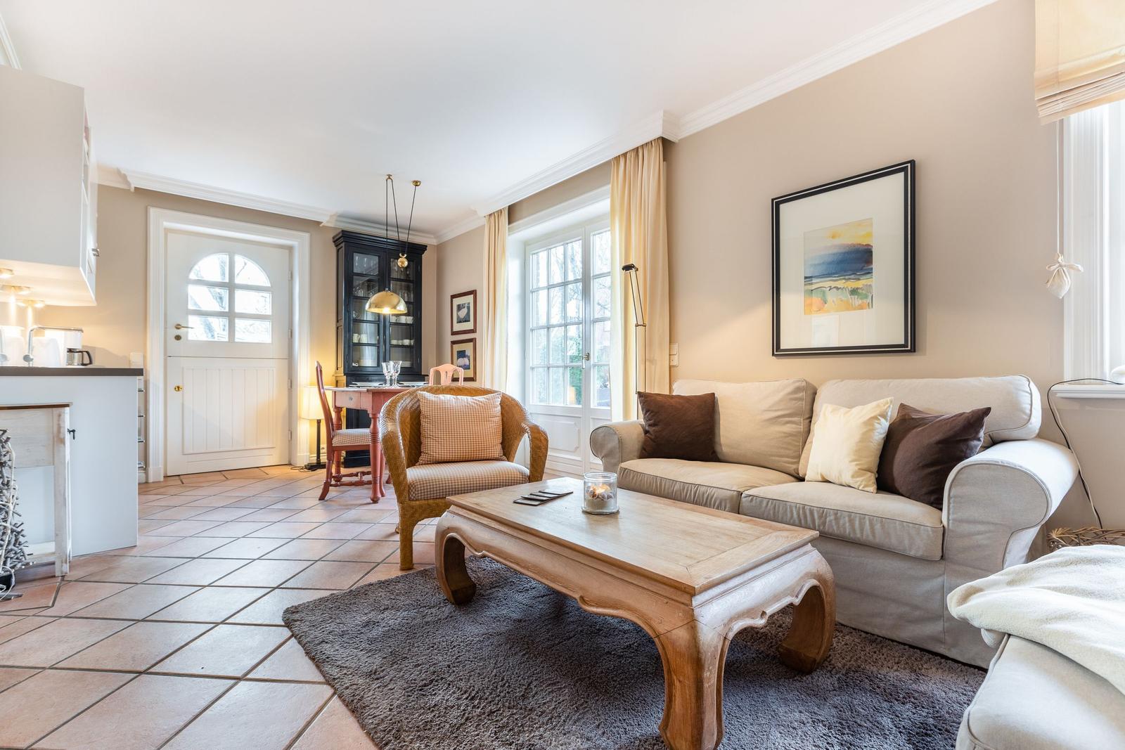 Living room with sofa, coffee table, and armchair. Windows, wall art, and kitchen area visible.