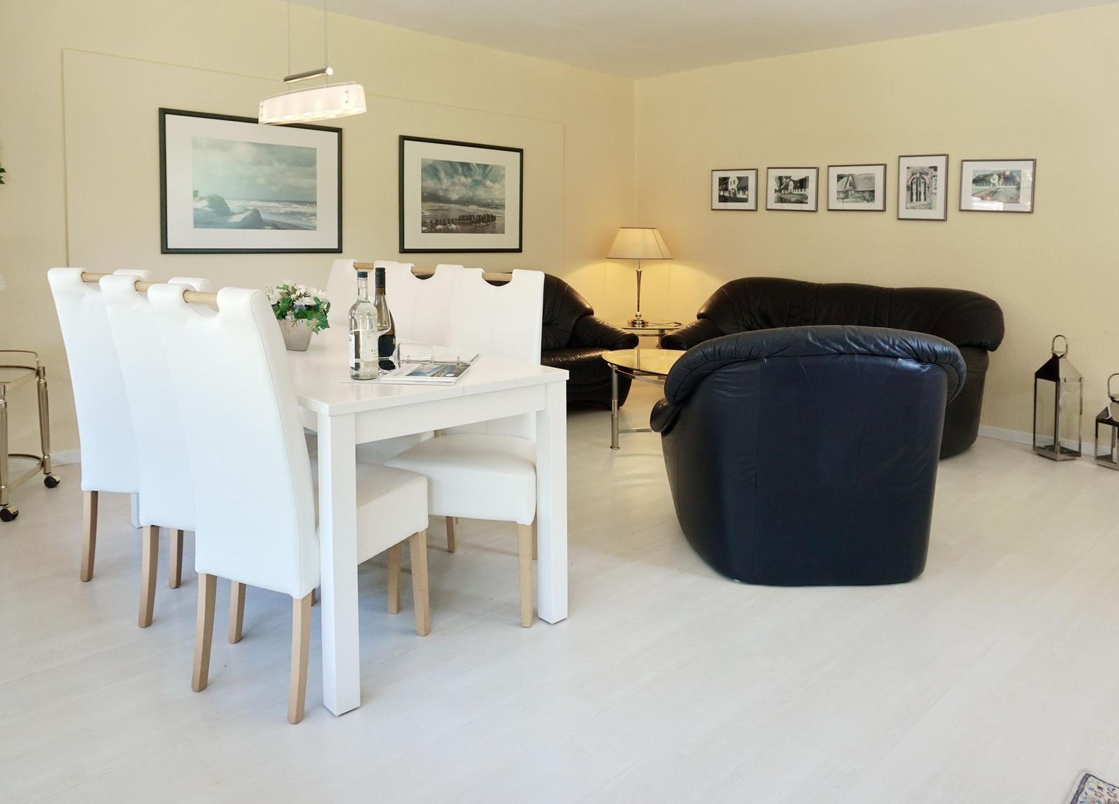 Dining area with white table and chairs. Living area with black leather chairs and wall pictures.