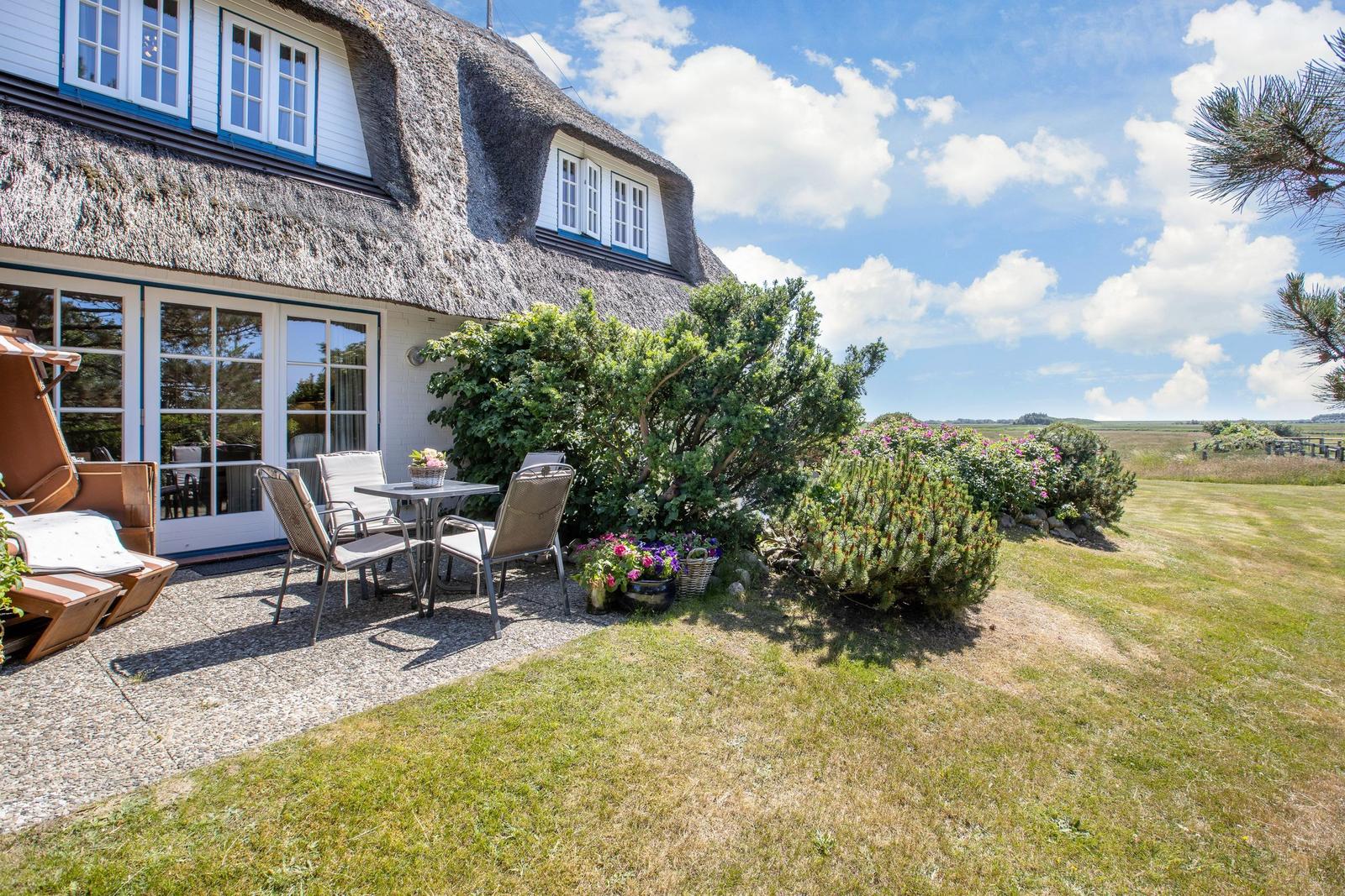 Haus mit Strohdach, Terrasse mit Tisch und Stühlen, Garten mit Blumen und Blick auf Felder.
