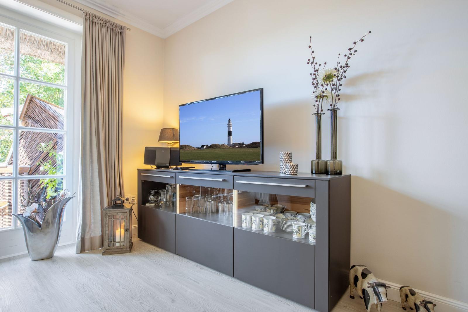 TV auf grauem Sideboard, Fenster mit Blick auf Garten, Dekoration mit Vasen und Kuhfiguren.