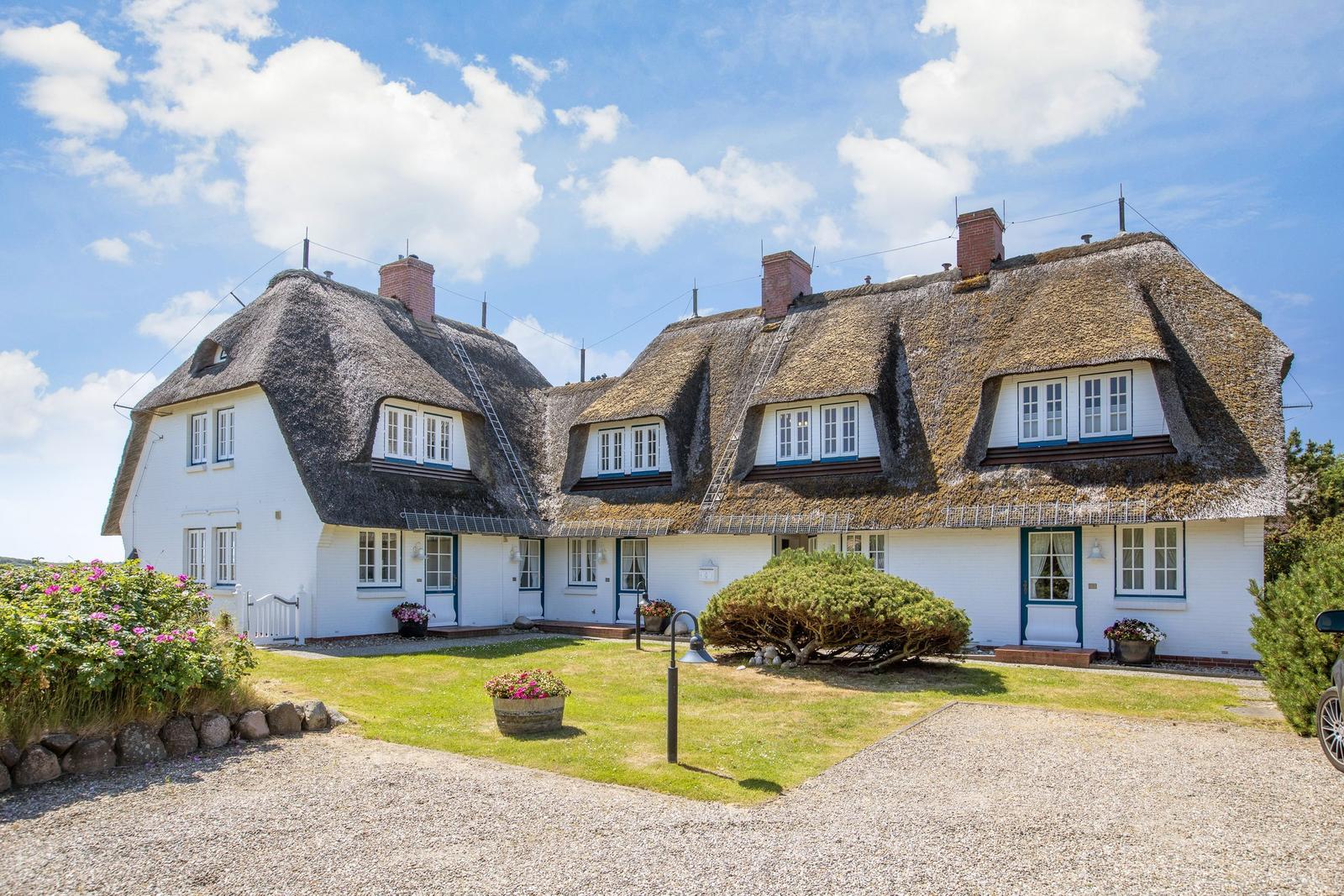 Traditionelles Haus mit Strohdach und weißer Fassade, umgeben von Grünfläche und Blumen.