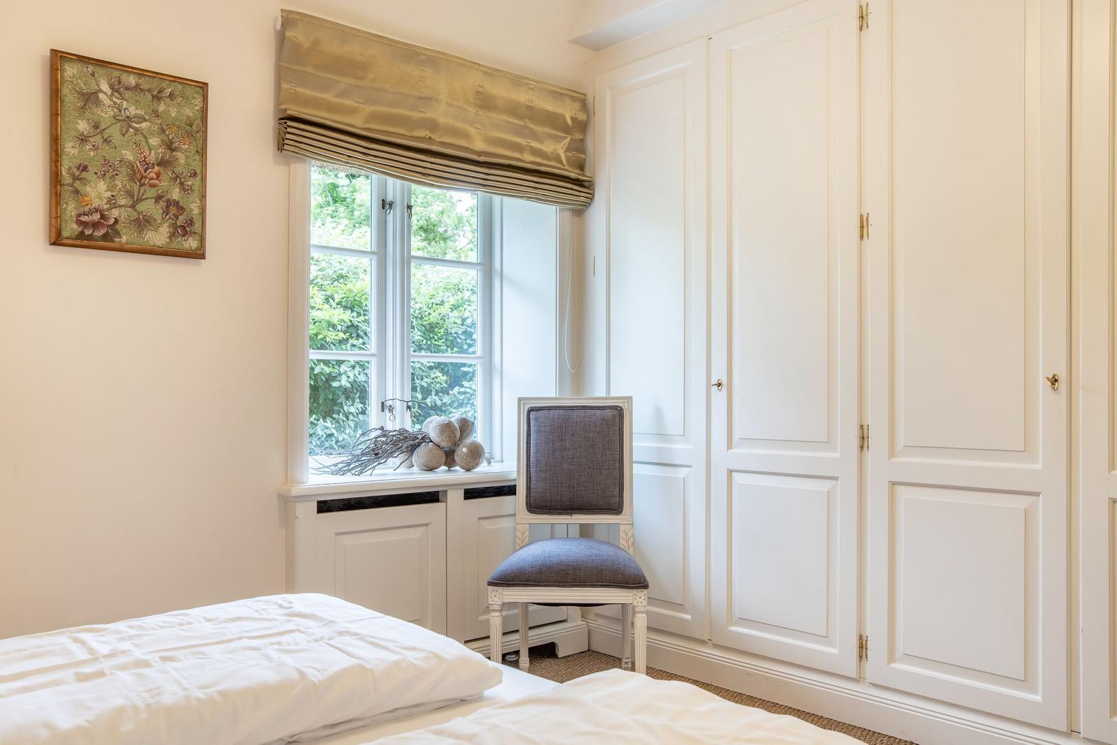 Bedroom with white bed, window, chair, and wardrobe.