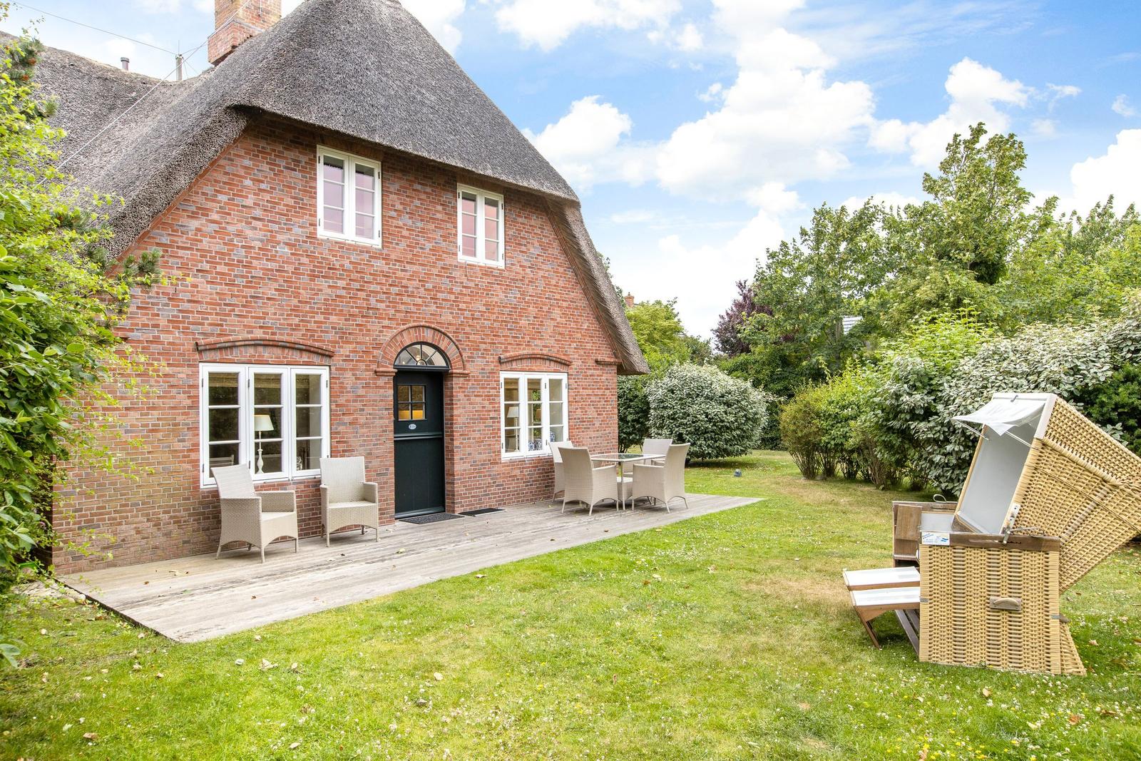 Red brick house with thatched roof, terrace, and garden with lounge chairs.