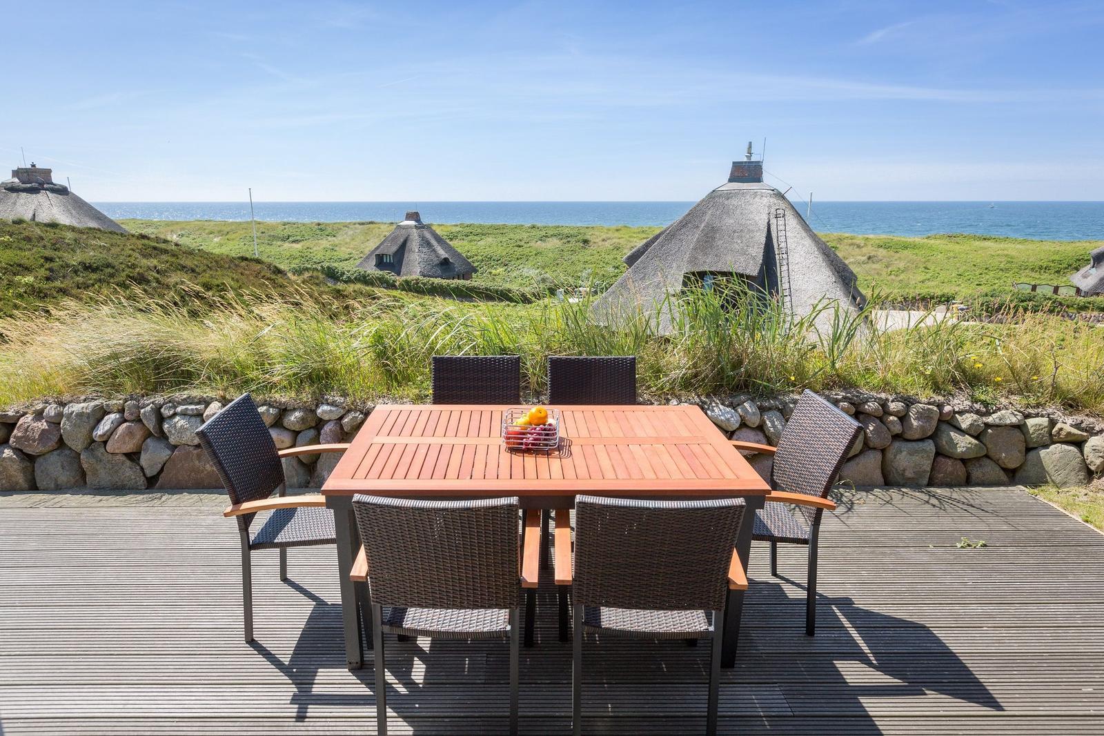 Terrasse mit Tisch und Stühlen, Blick auf Strand und Dachhäuser.