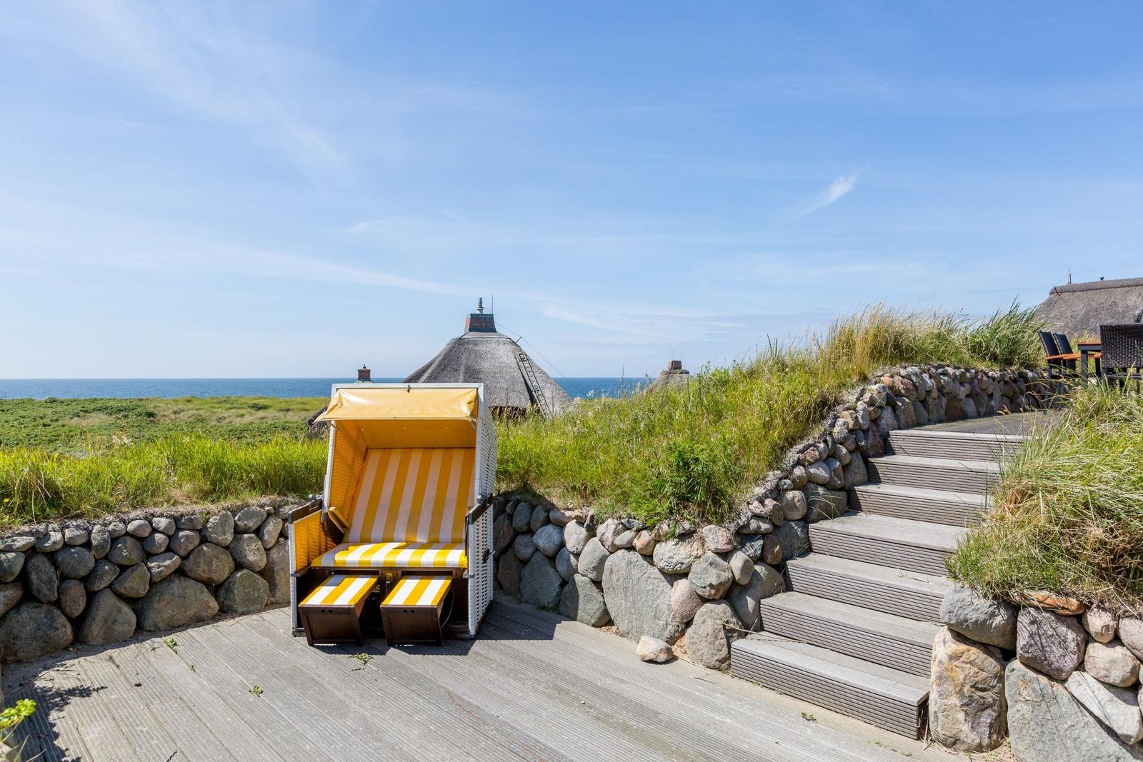 Ein gelber Strandkorb steht auf einer Holzterrasse mit Steintreppe und Blick auf das Meer.