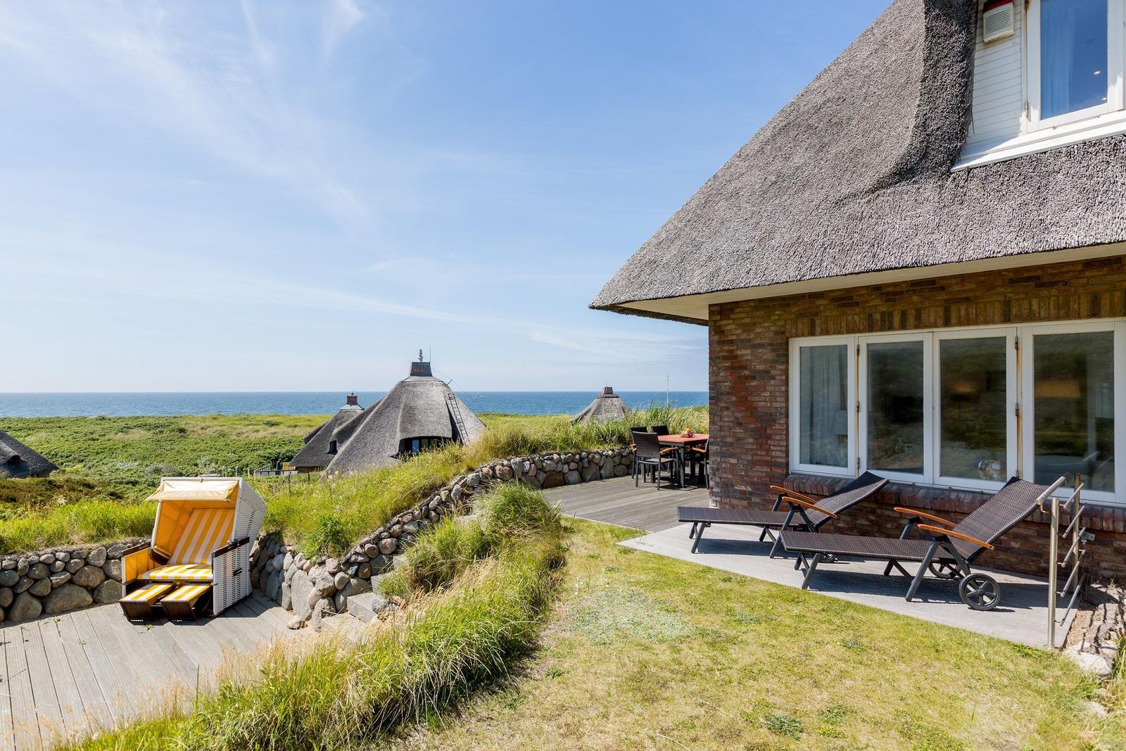 Haus mit Dach aus Stroh, Terrasse mit Liegen und Blick auf das Meer.
