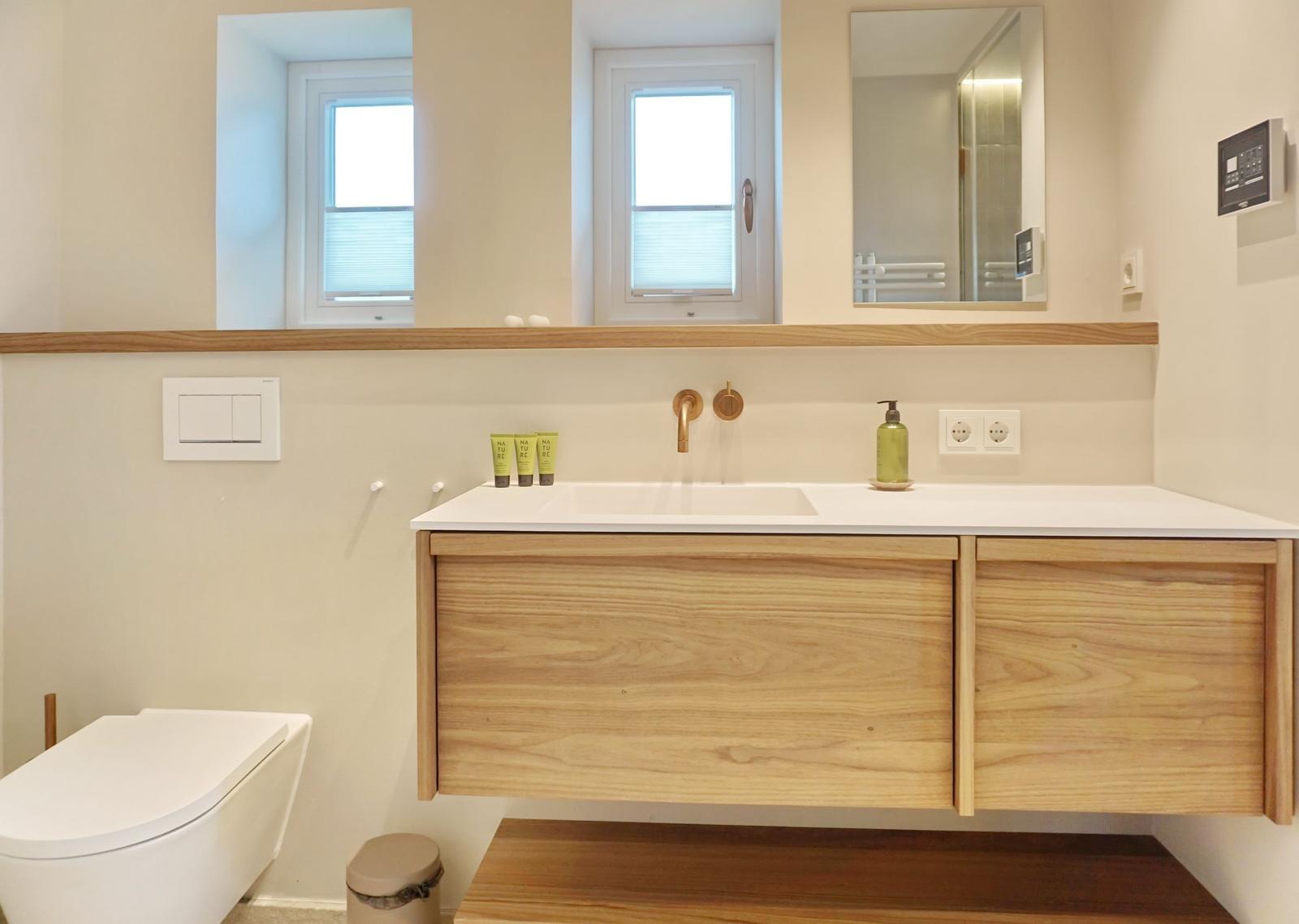 Bad mit Waschbecken, Spiegel und Toilette. Holzunterschrank und Fenster.