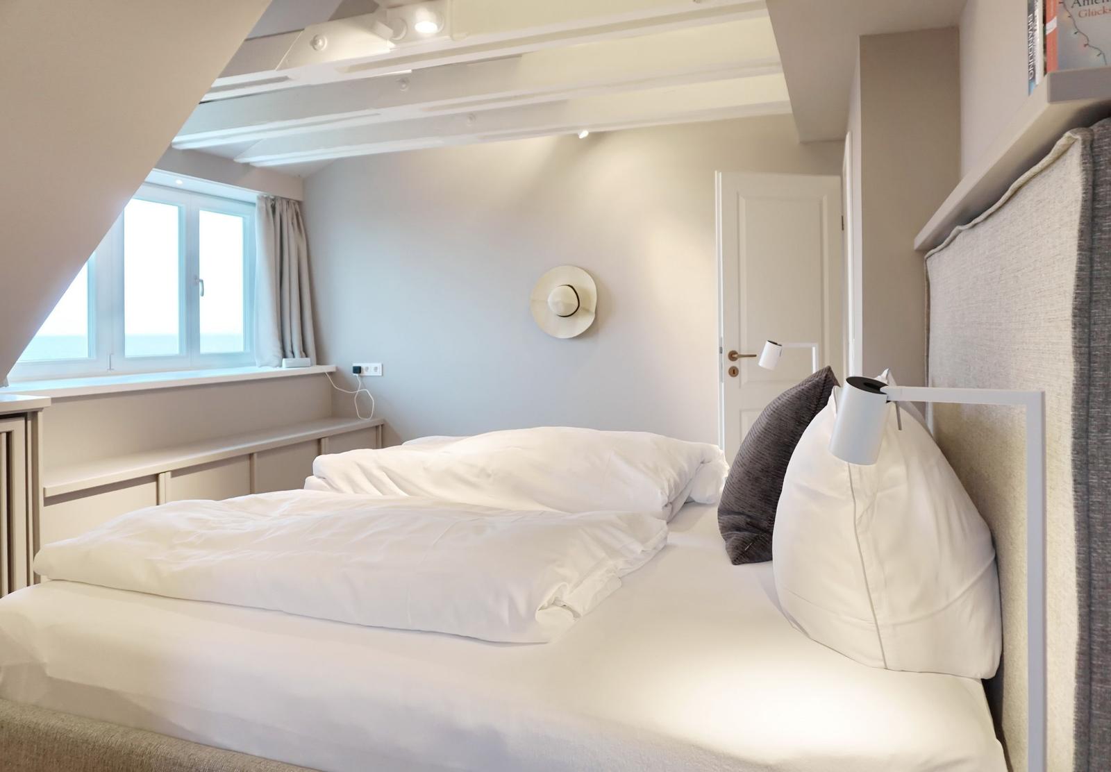 Schlafzimmer mit Dachschrägen, Fenster und Bett mit weißen Laken.