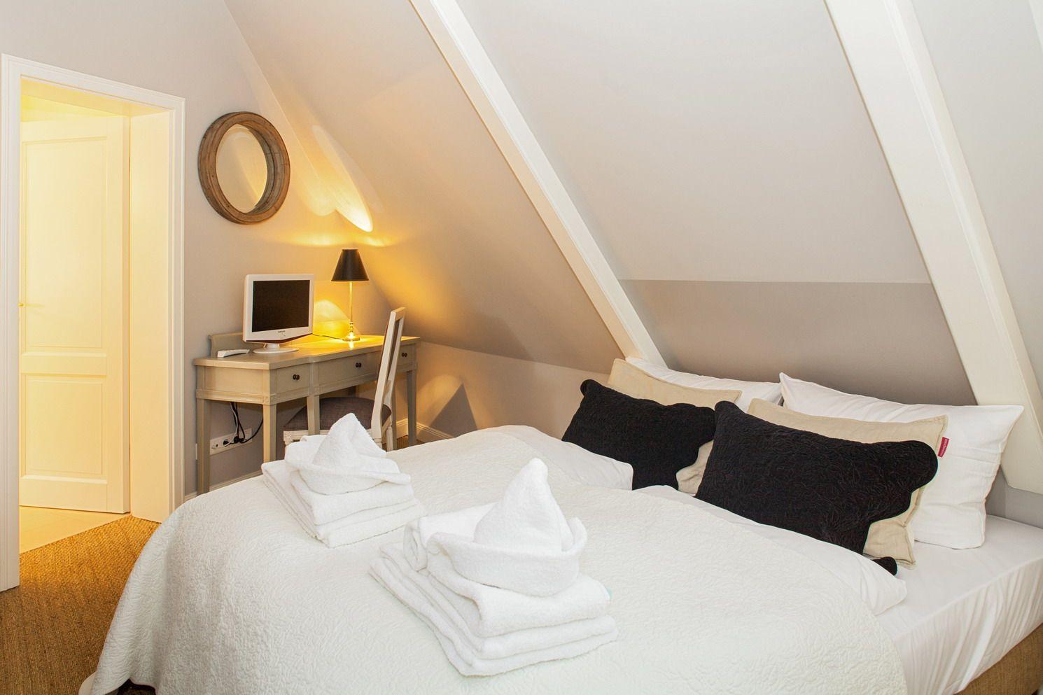 Schlafzimmer mit Dachschräge, Bett mit Kissen und Handtüchern, Schreibtisch mit Monitor und Lampe.
