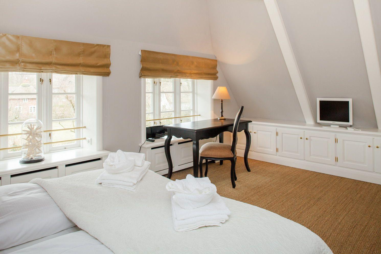 Schlafzimmer mit Dachschräge, zwei Fenstern, Schreibtisch und Bett mit Handtüchern.