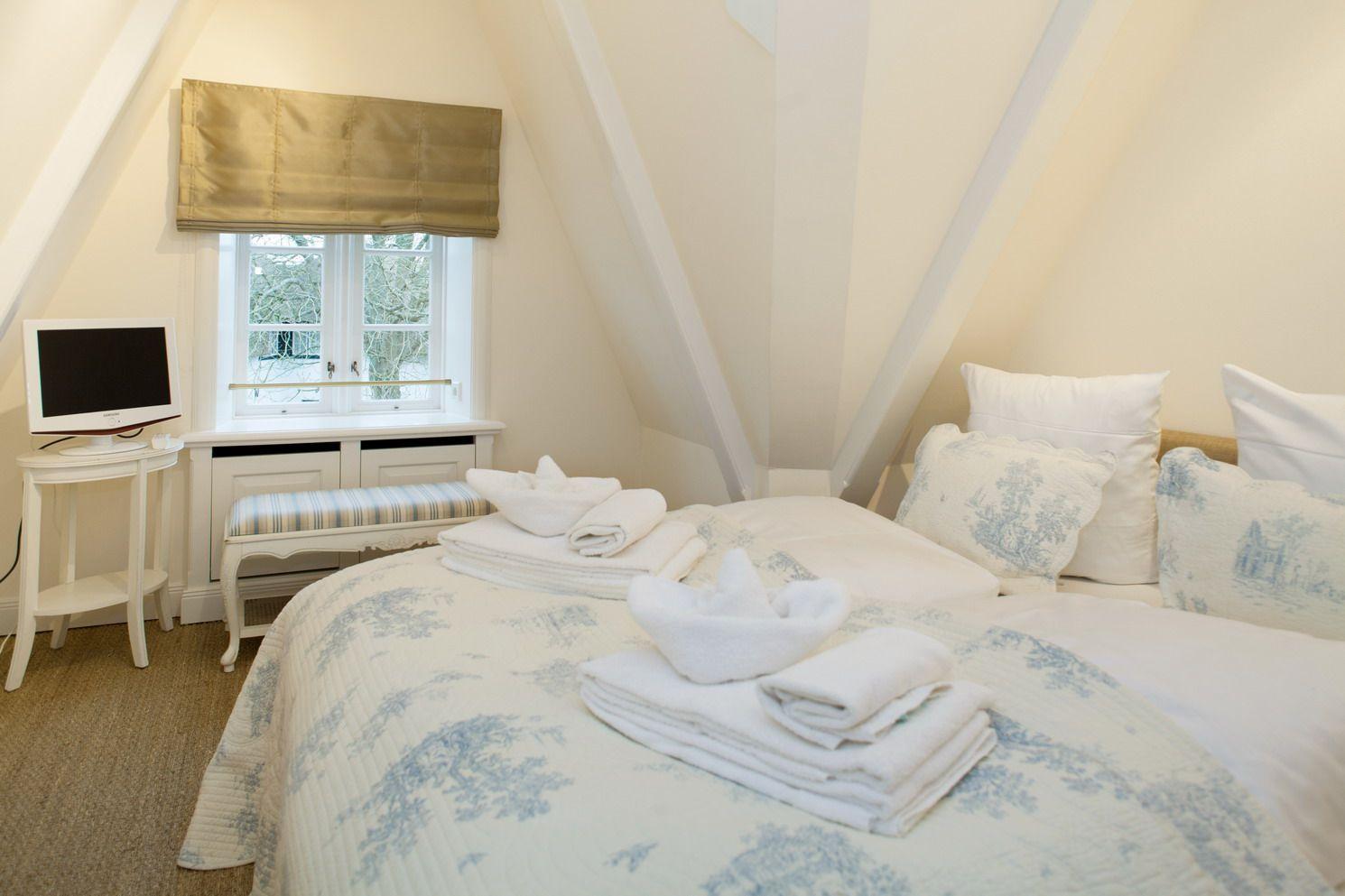 Bedroom with sloped ceiling, bed with blue and white bedding, and window with gold roman shades.