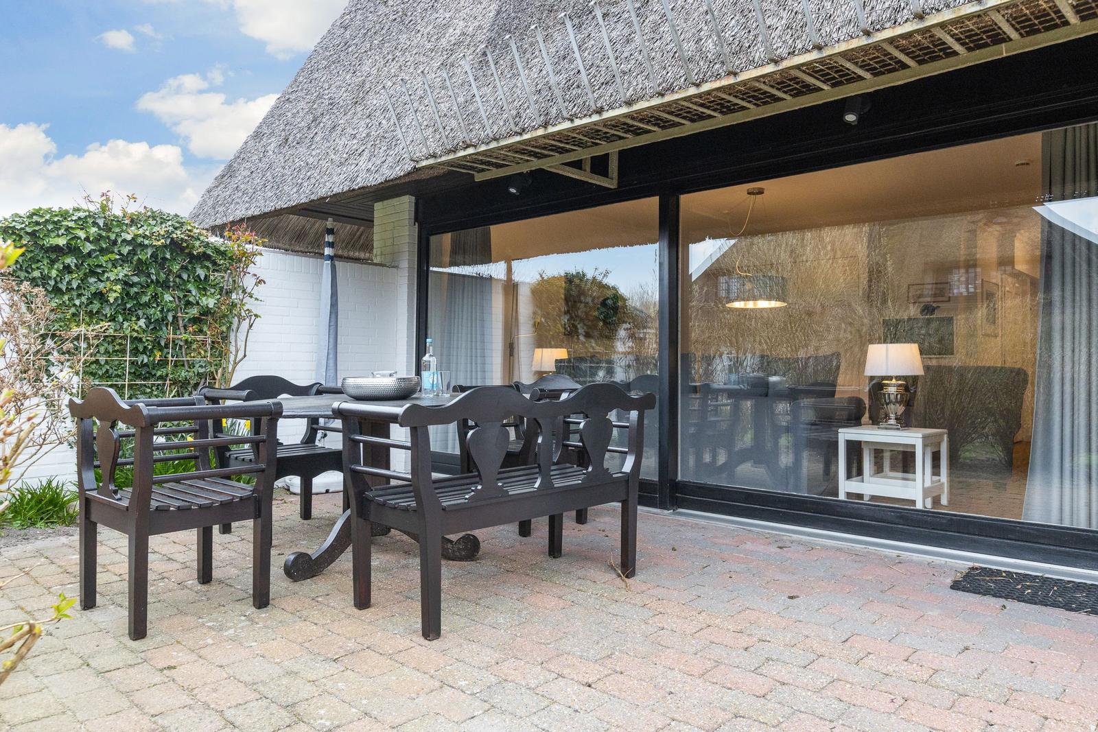 Terrasse mit Tisch und Stühlen unter strohgedecktem Dach. Große Glasfront zeigt Innenraum.