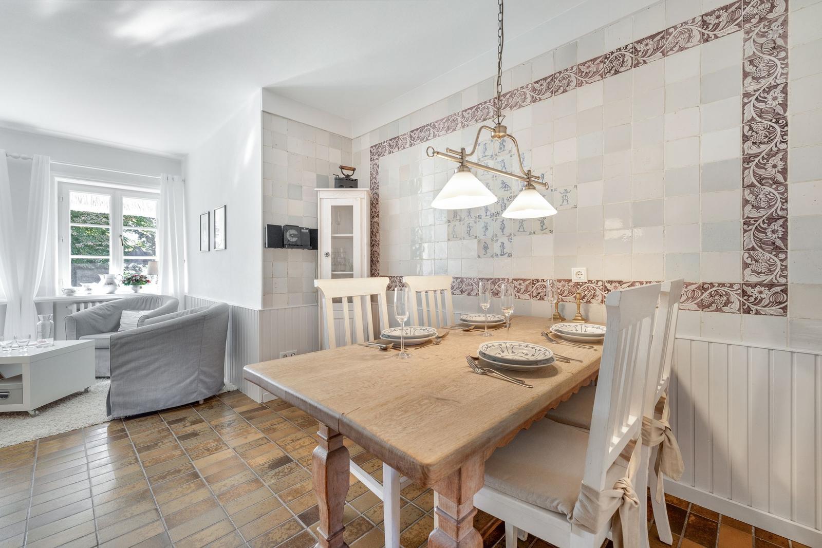 Dining room with wooden table, white chairs, and decorative tiled walls. Living area with sofa and window.