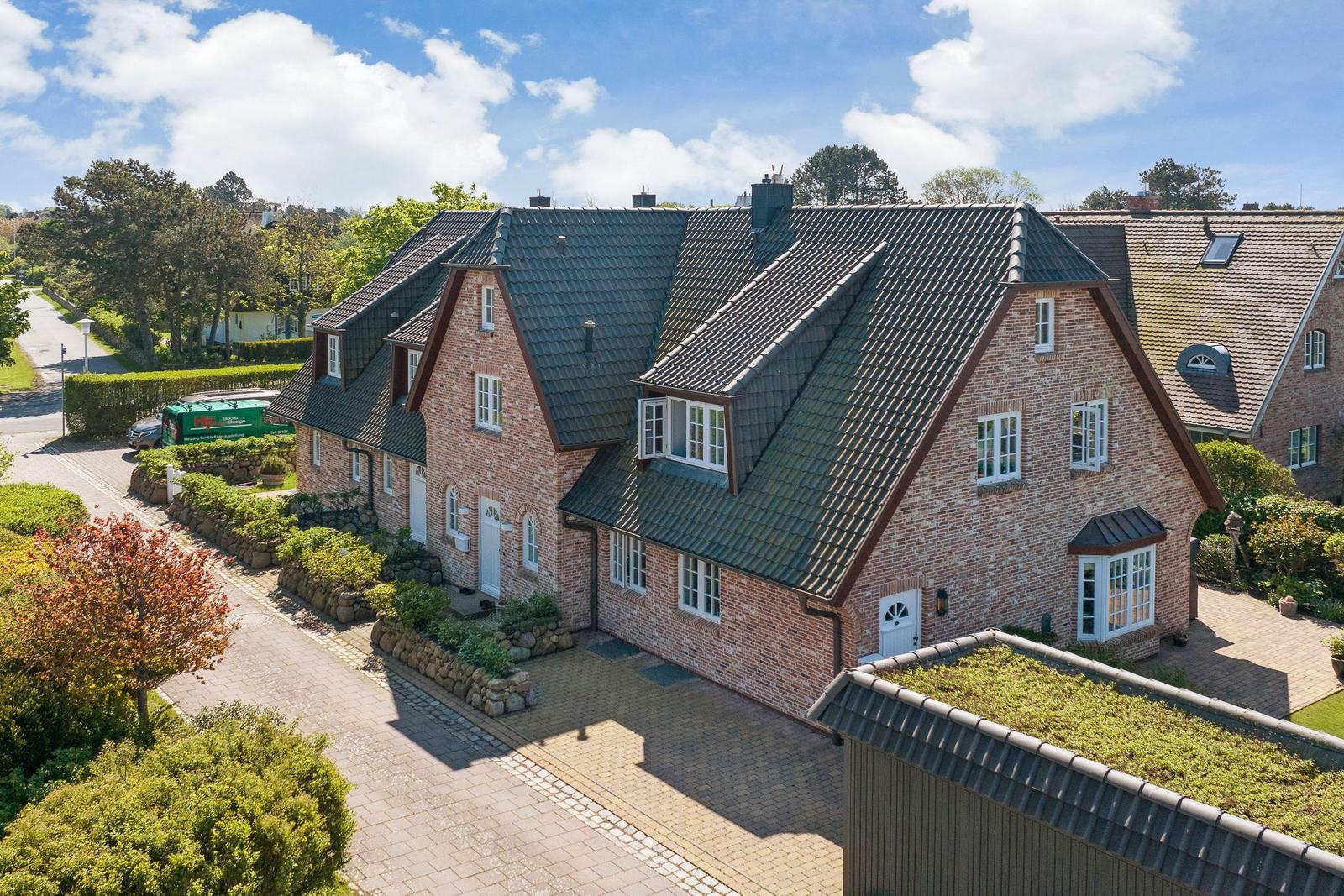 Haus mit Ziegelfassade und grünem Dach, umgeben von gepflegtem Garten und Steinmauern.