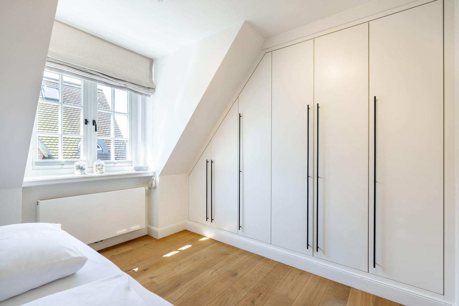 Schlafzimmer mit Dachschrägen, weißem Schrank und Holzboden. Fenster mit Blick auf Dächer.