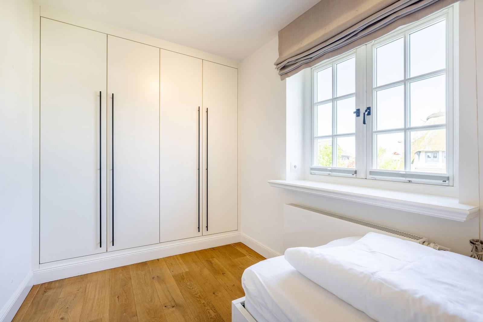 Schlafzimmer mit weißem Schrank, Holzboden und Fenster mit Blick nach draußen.