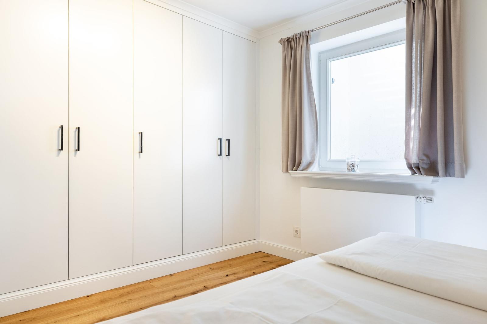 Schlafzimmer mit weißem Schrank, Holzboden und Fenster mit Vorhängen.