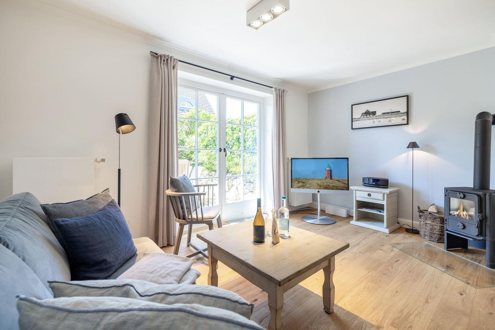 Wohnzimmer mit Sofa, Couchtisch, Fernseher und Kaminofen. Fenster mit Blick auf Garten.