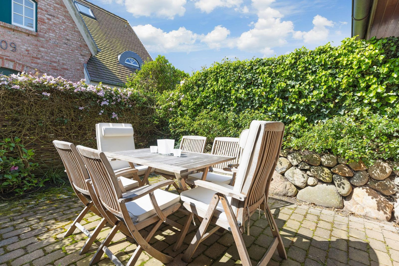 Terrasse mit Holztisch und sechs Stühlen, umgeben von grüner Hecke und Steinmauer.