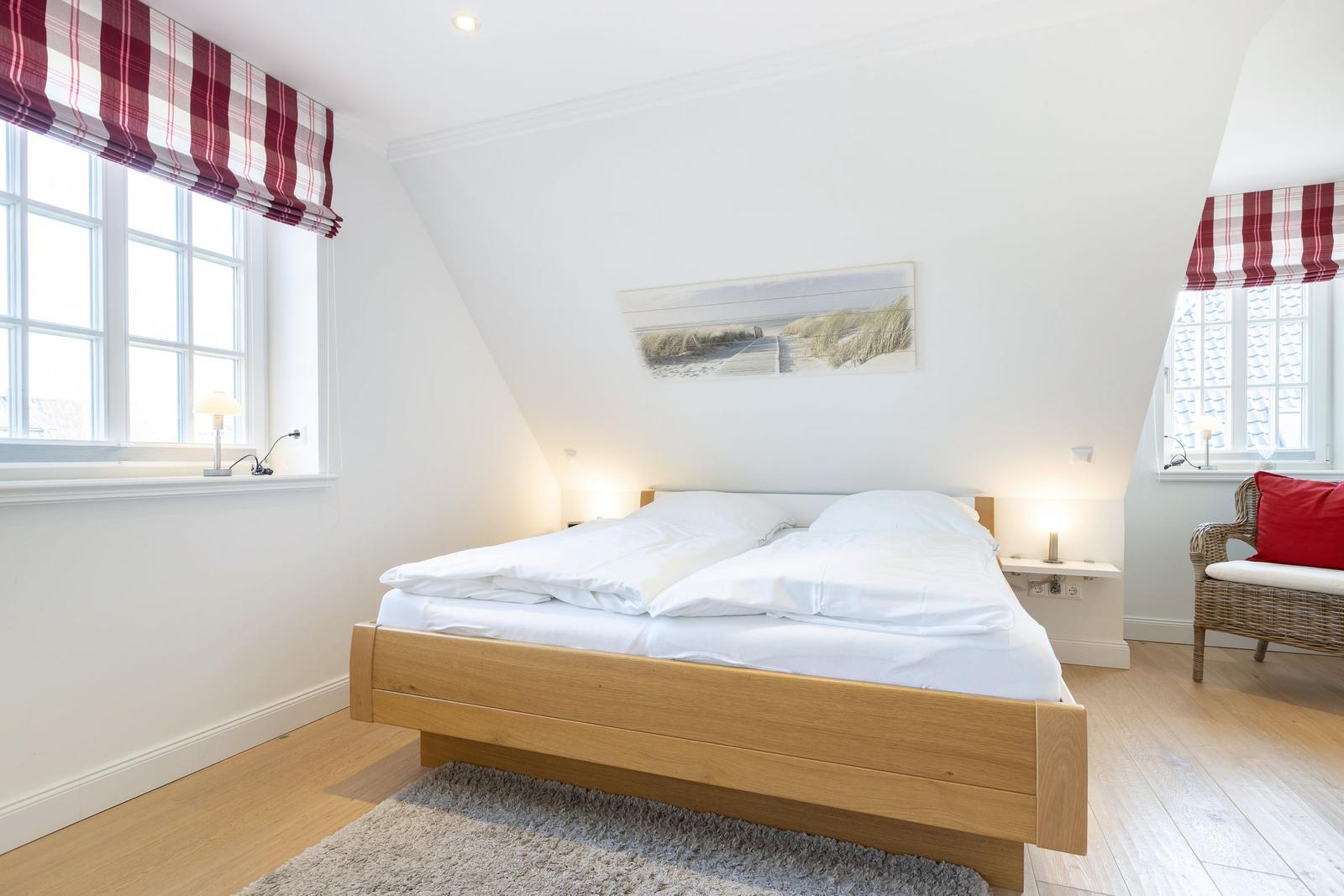 Schlafzimmer mit Doppelbett, Holzboden und zwei Fenstern mit roten Gardinen.