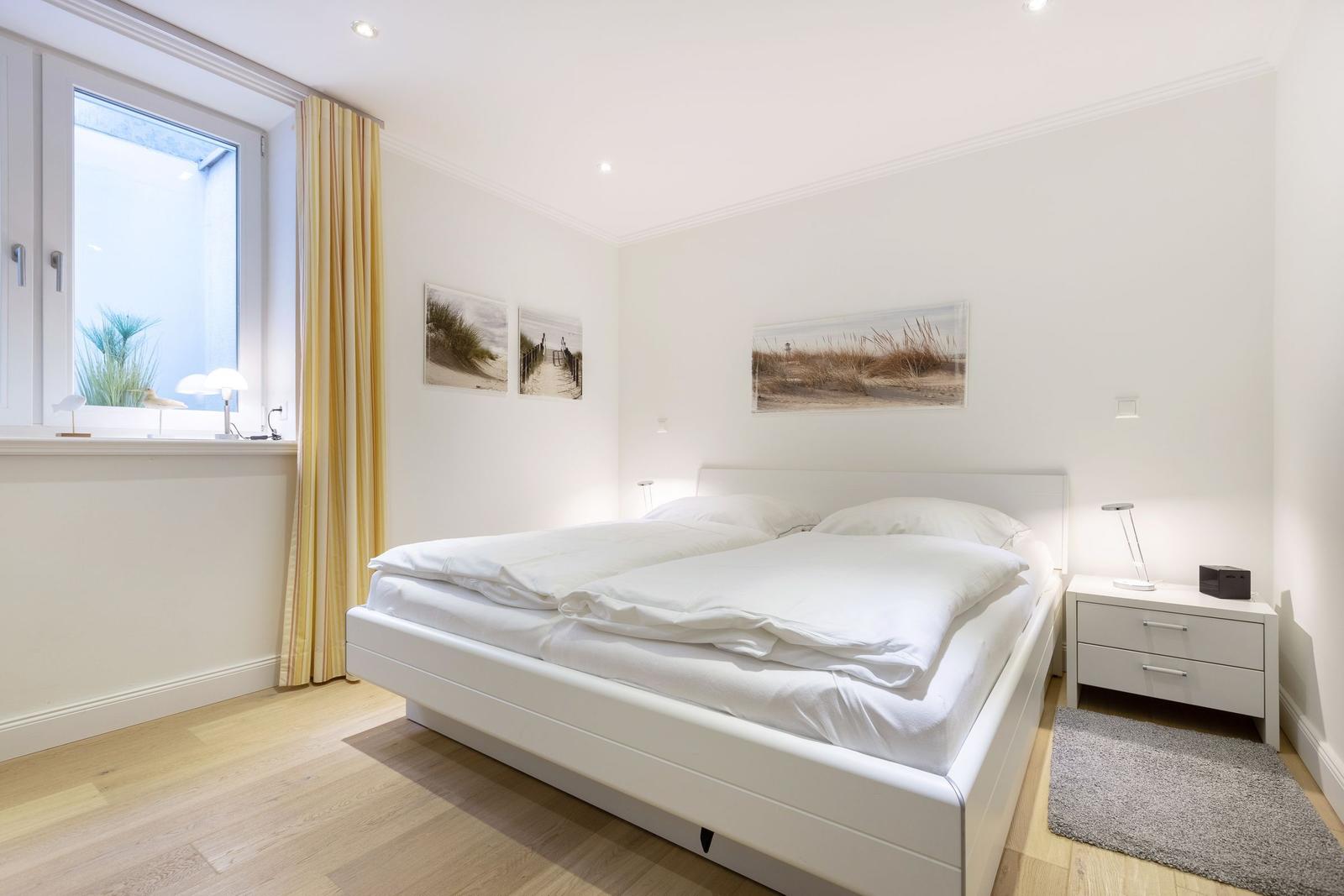 Schlafzimmer mit weißem Bett, Holzboden und Fenster mit gelben Vorhängen.