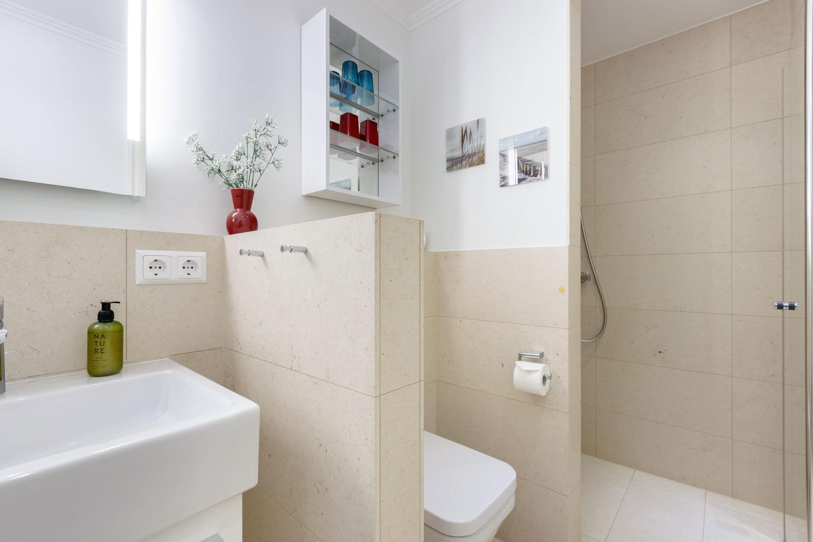 Bad mit Dusche, Waschbecken und Toilette. Wandregal mit Gläsern und Blumen.