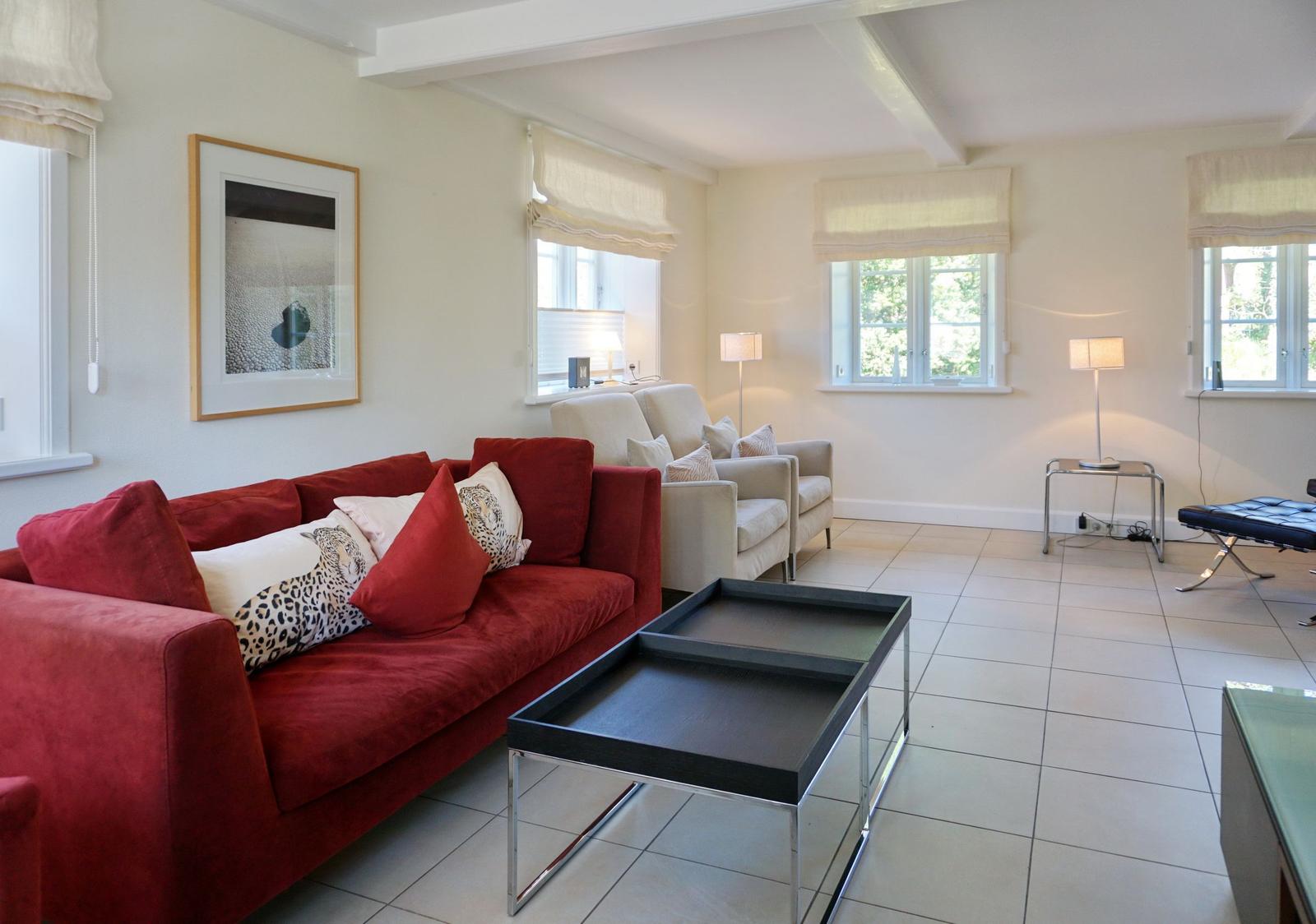 Ein Wohnzimmer mit rotem Sofa, weißen Sesseln und Fenstern mit Rollläden.