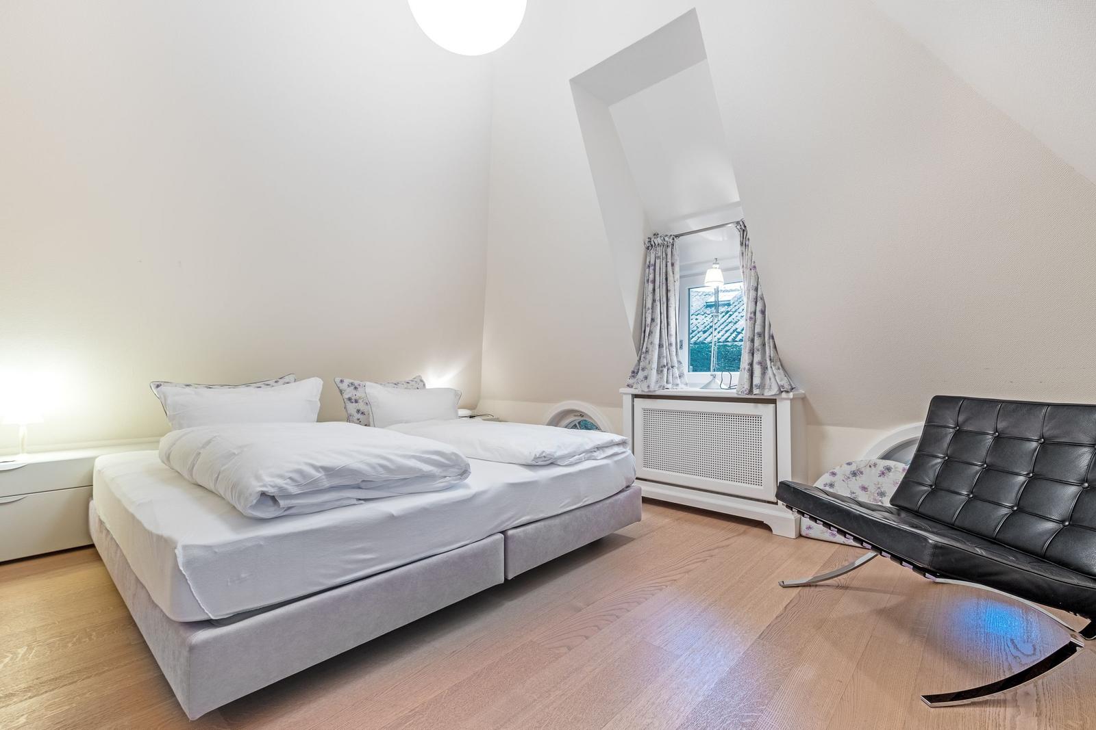 Schlafzimmer mit Doppelbett, schwarzer Ledersessel und schräger Fensterfront.