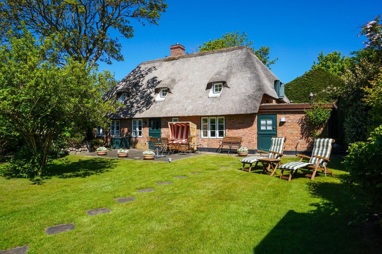 Haus mit Strohdach, Garten mit Liegestühlen und Sonnenschirm.