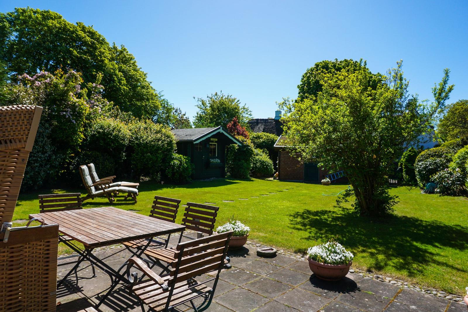Garten mit Terrasse, Holztisch, Stühlen und Blumenkübeln. Grasfläche und Bäume im Hintergrund.