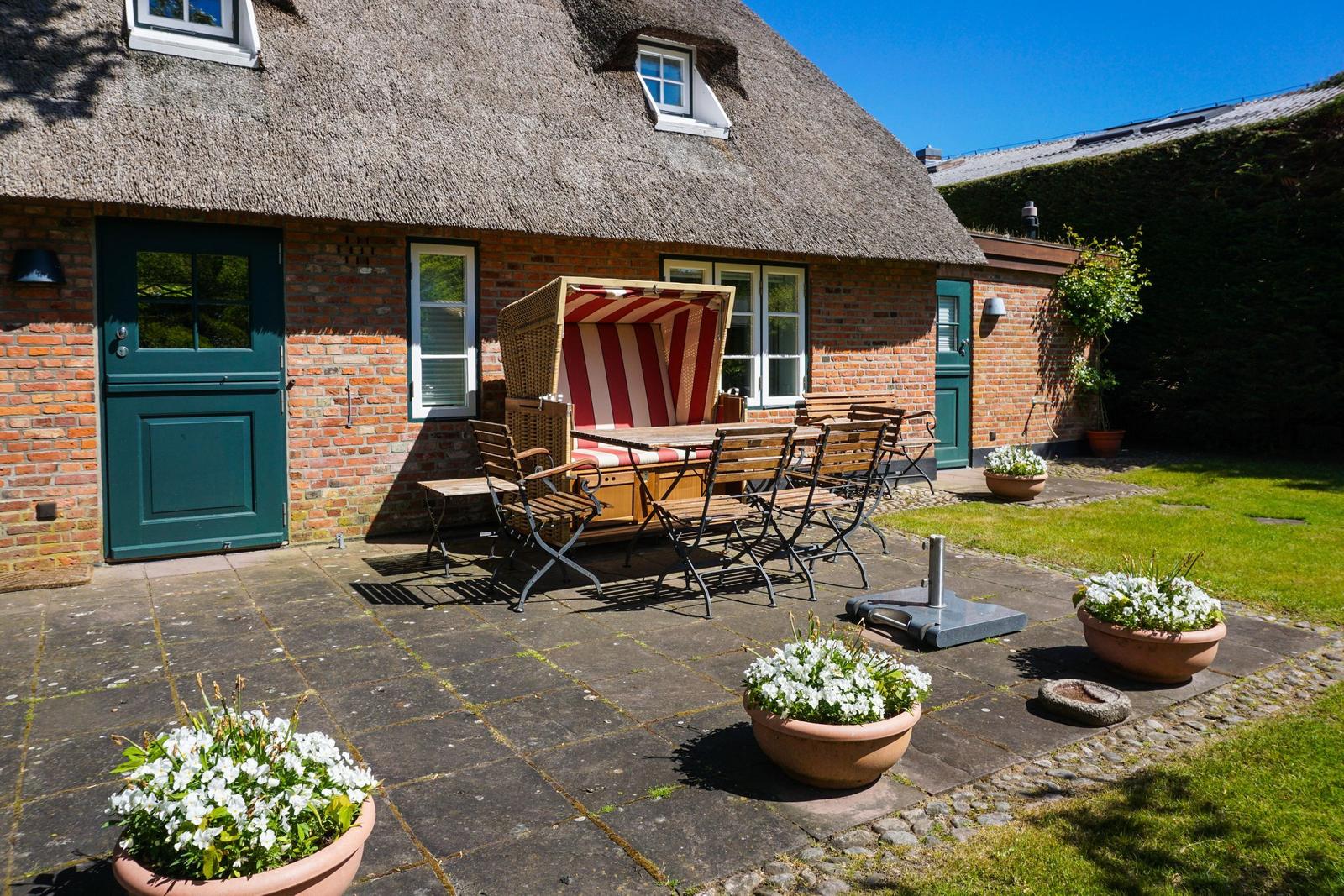 Haus mit strohgedecktem Dach, Garten mit Tisch, Stühlen und Blumenkübeln.