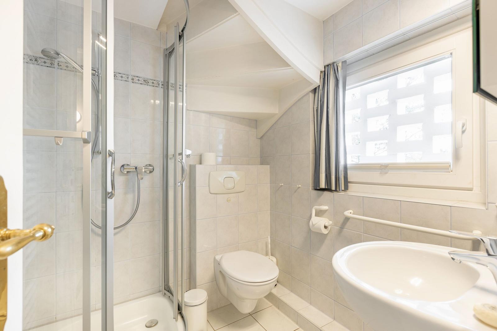 Badezimmer mit Dusche, Toilette und Waschbecken. Fenster mit Vorhang.