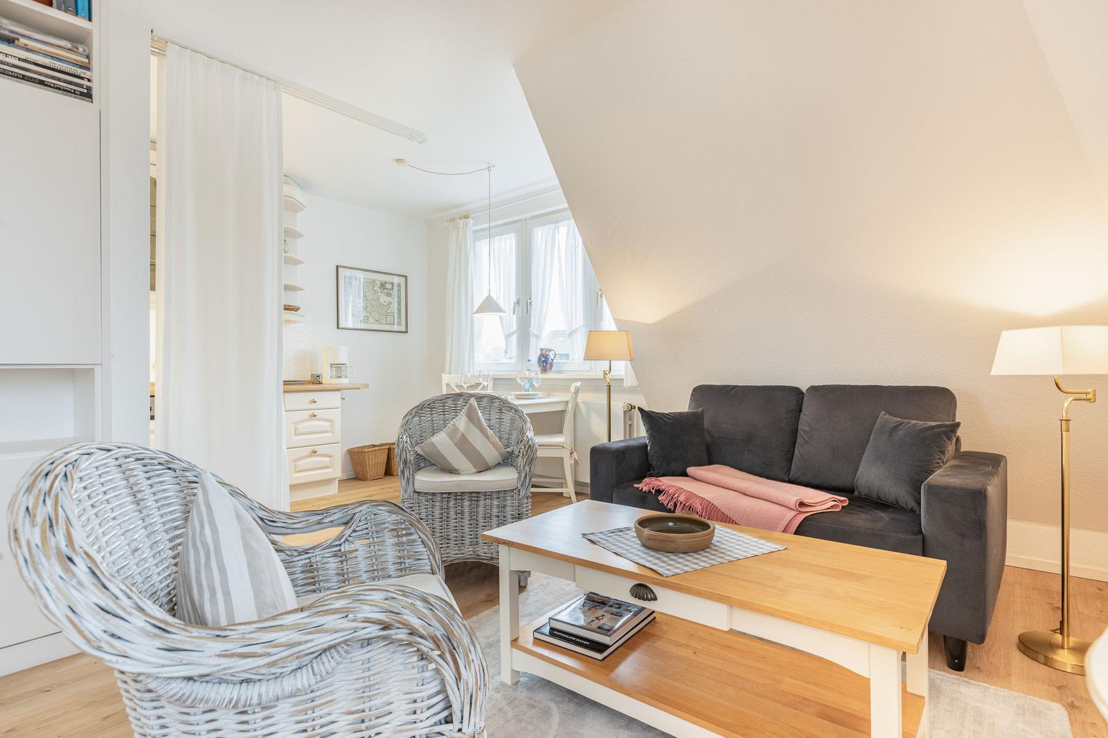 Wohnzimmer mit grauem Sofa, weißen Rattan-Stühlen und Holztisch. Dachschrägen und Fenster sichtbar.