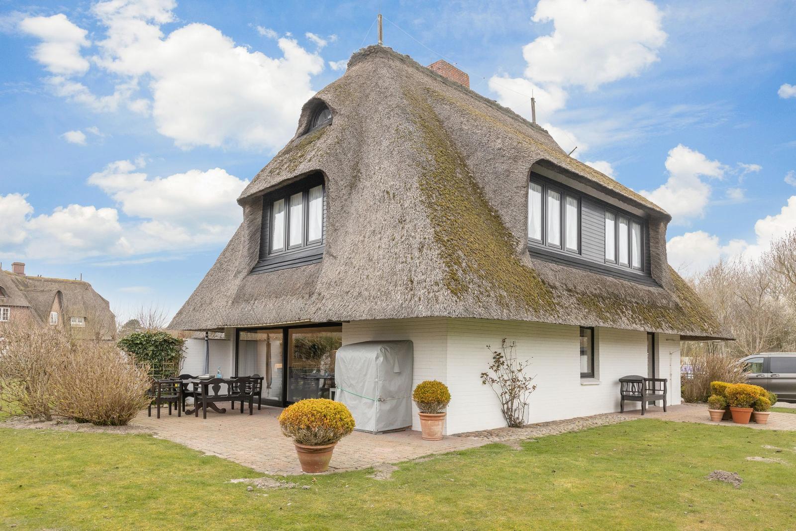 Haus mit Strohdach, Terrasse und Garten unter blauem Himmel.