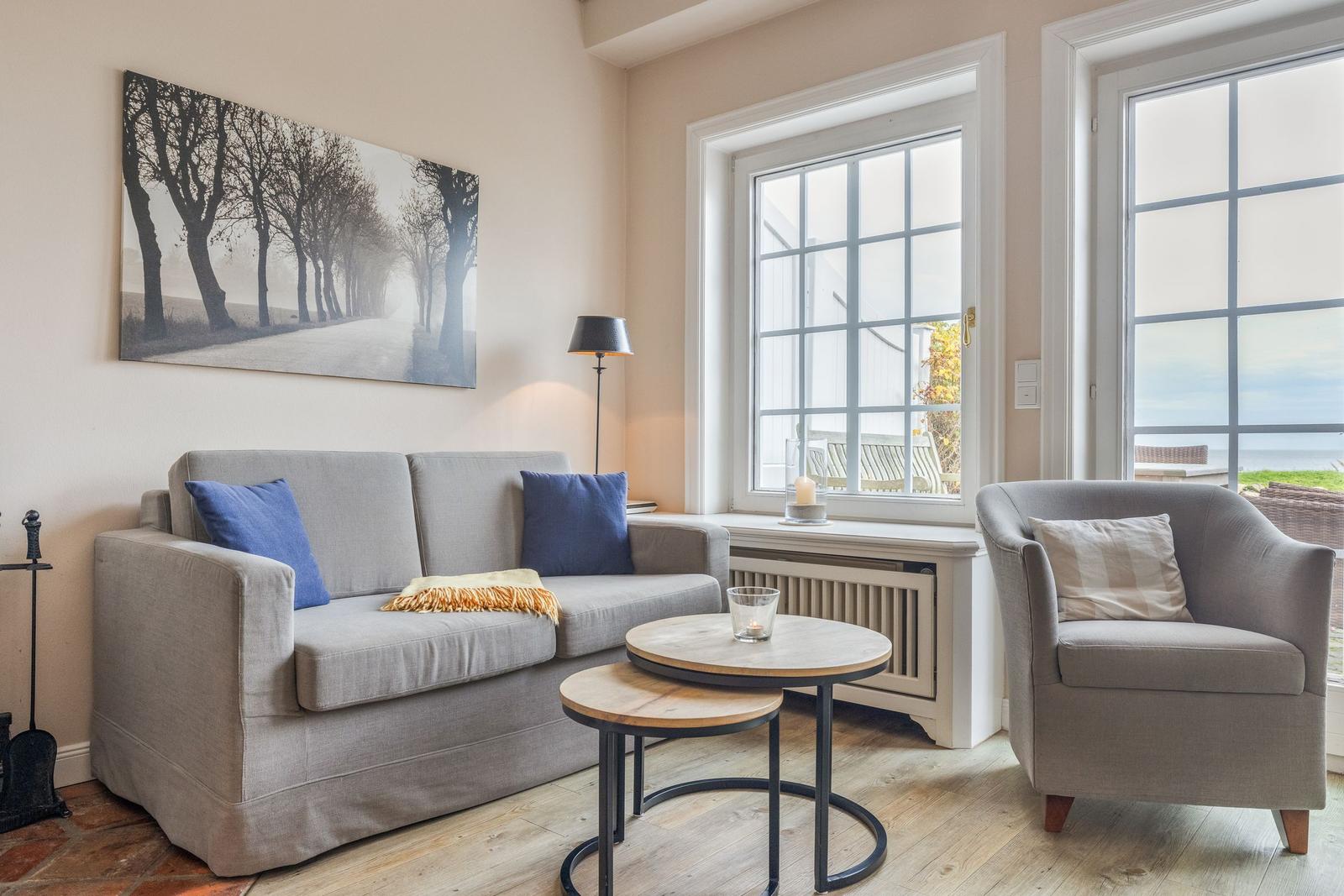 Ein Wohnzimmer mit Sofa, Sessel und Tisch. An der Wand hängt ein Bild. Durch Fenster ist die Aussicht aufs Meer.