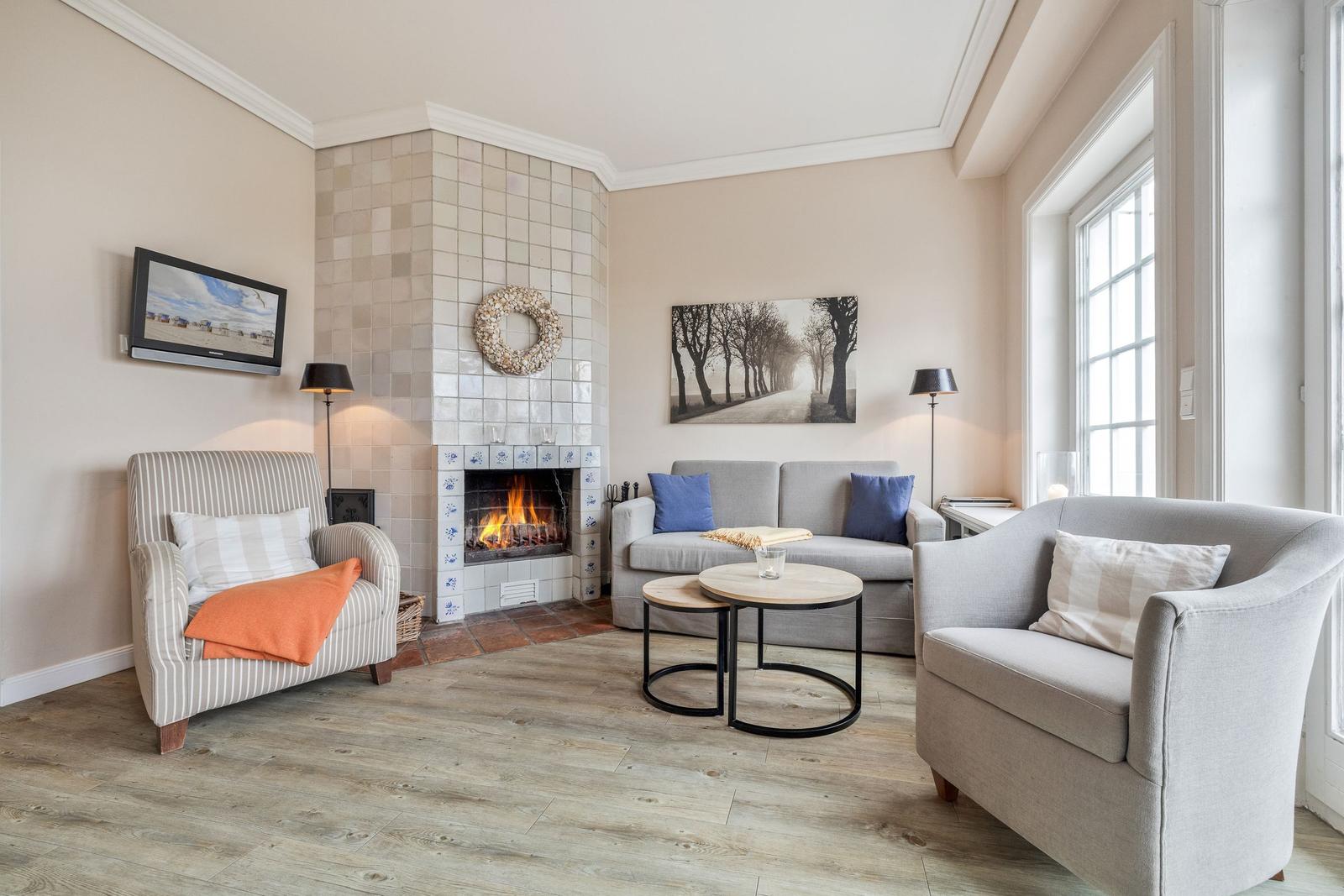 Wohnzimmer mit Kamin, Sofa und zwei Sesseln. Holzboden und große Fenster.