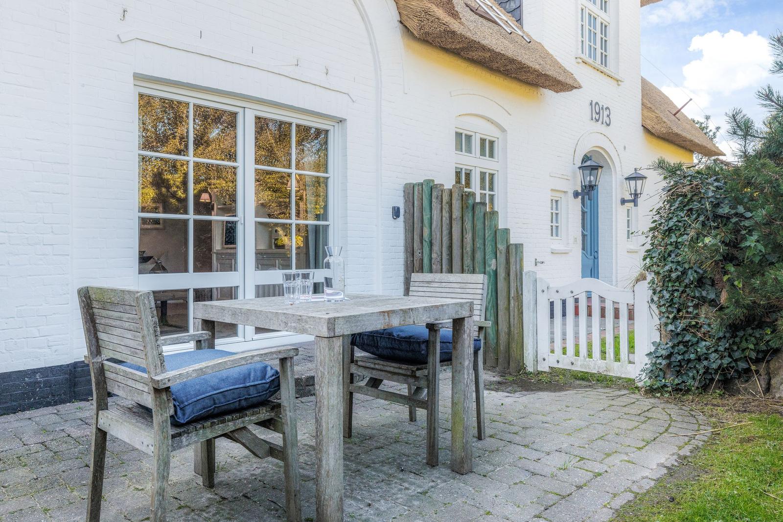 Wooden table with chairs on paved courtyard in front of white house with thatched roof.