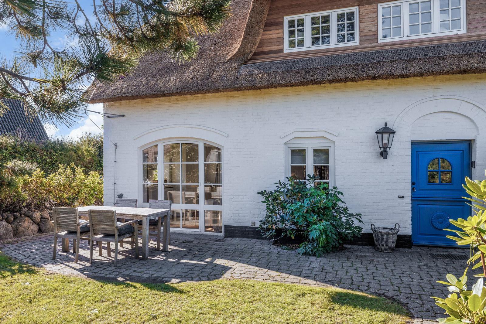 Weiße Hauswand mit blauer Tür und strohgedecktem Dach. Außenterrasse mit Tisch und Stühlen.