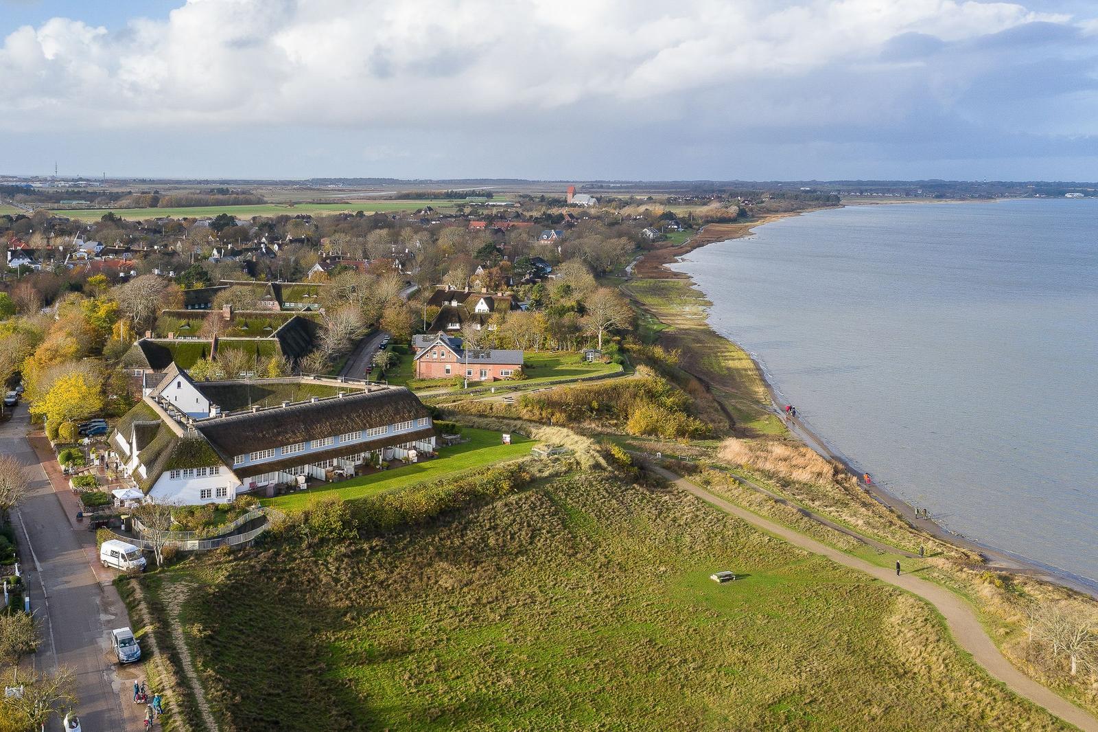 Haus mit Dachrinne am Wasser, Grünflächen und Wanderweg entlang der Küste.