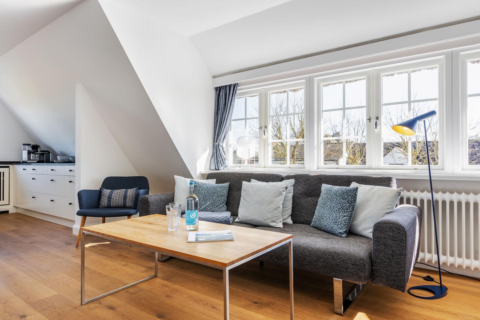 Wohnzimmer mit Sofa, Couchtisch und Fenstern. Holzboden und weiße Wände.