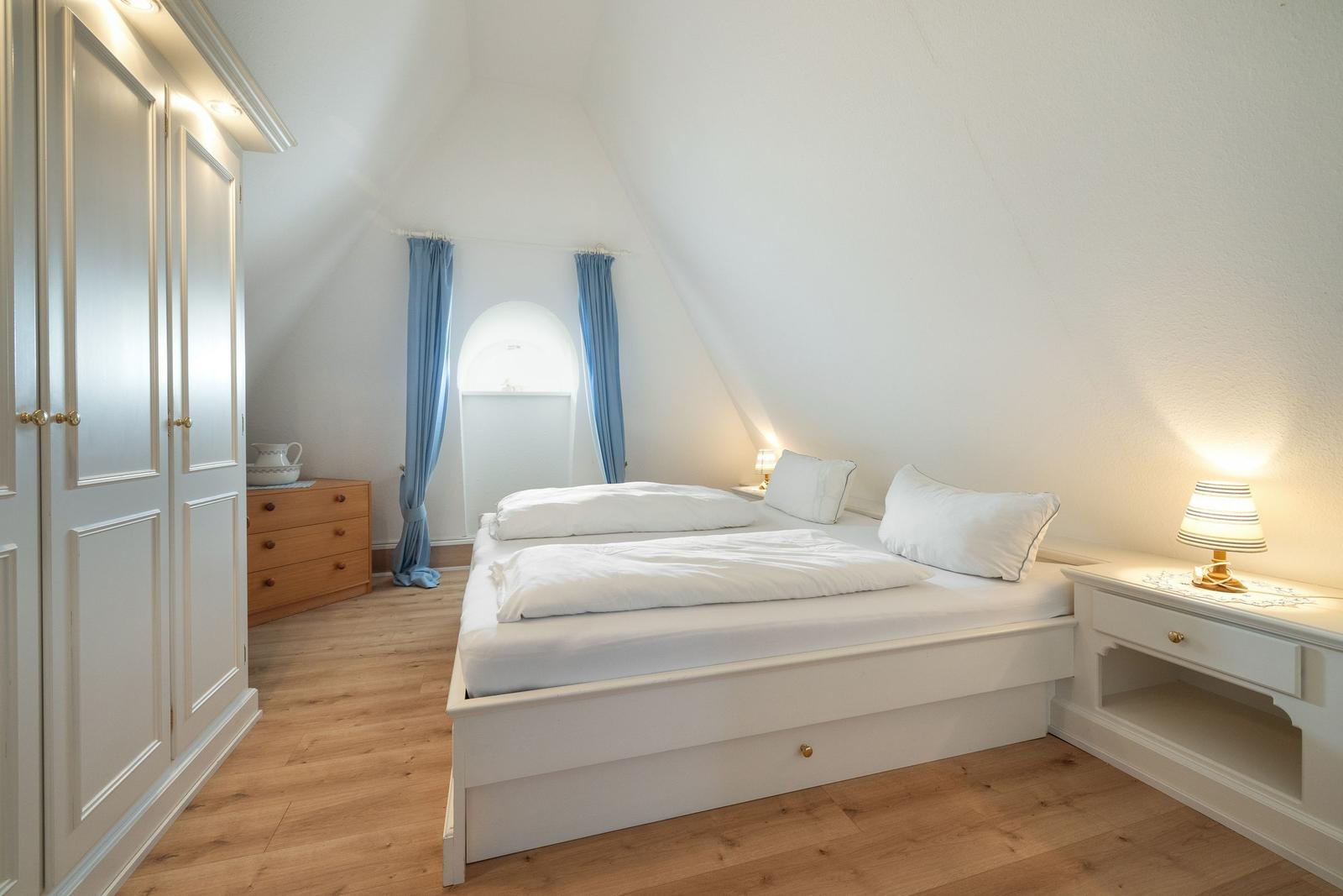 Schlafzimmer mit weißem Bett, Holzboden und Schrank. Fenster mit blauen Vorhängen.