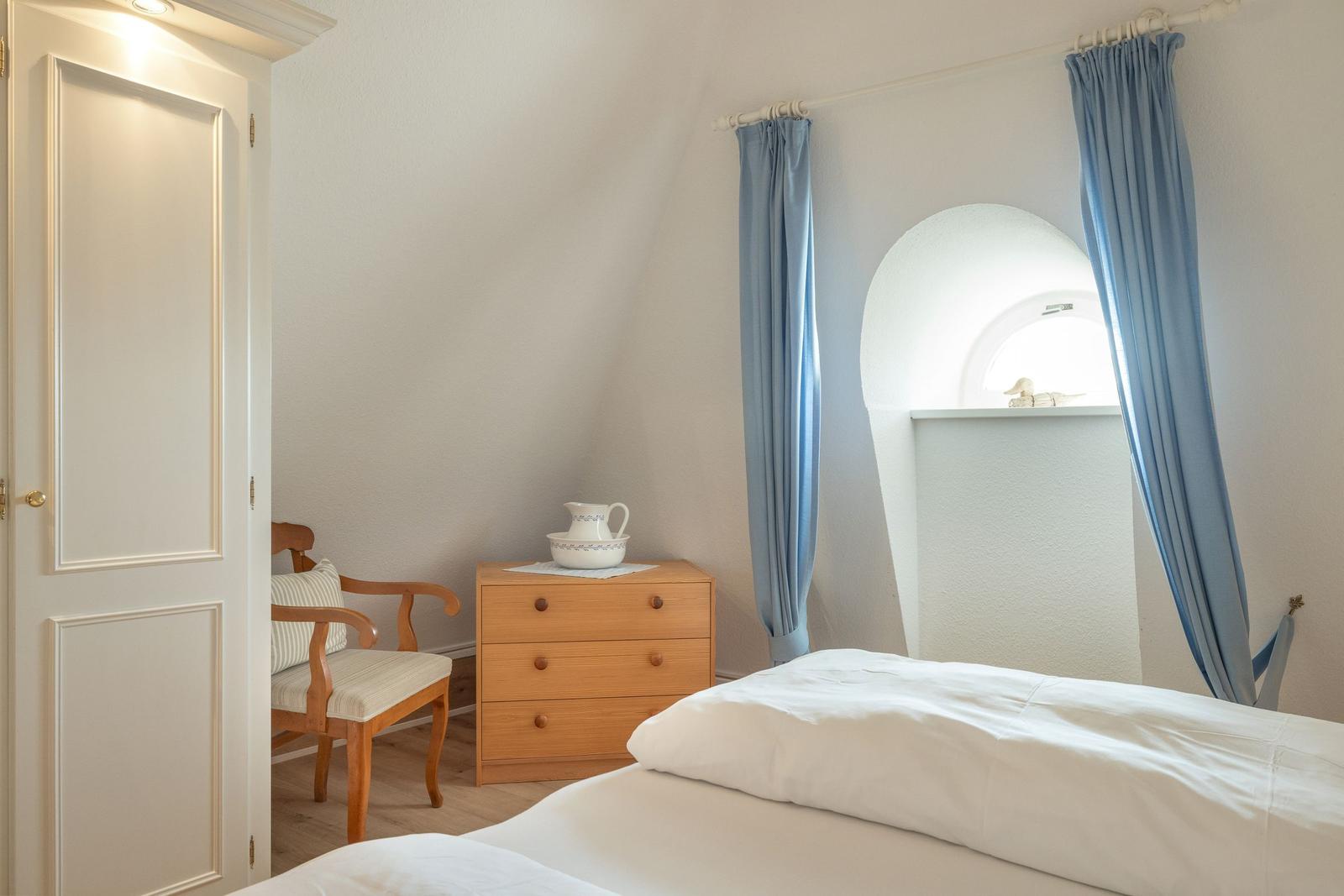 Schlafzimmer mit Bett, Schrank und Stuhl. Fenster mit blauen Vorhängen.