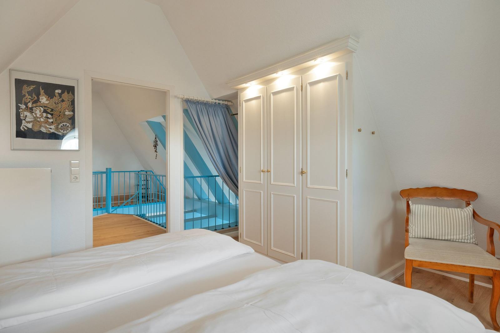 Schlafzimmer mit Bett, Kleiderschrank und Stuhl. Blick in einen Flur mit blauer Brüstung.