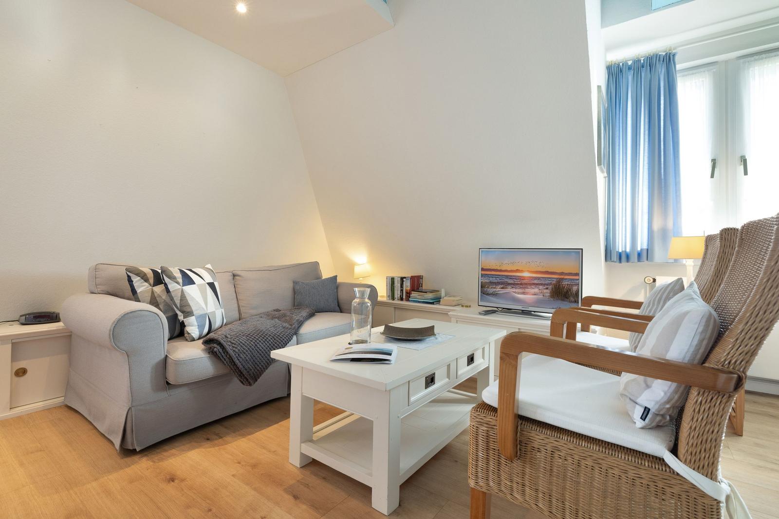 Wohnzimmer mit Sofa, zwei Stühlen und Fernseher. Holzboden und Fenster mit blauen Vorhängen.