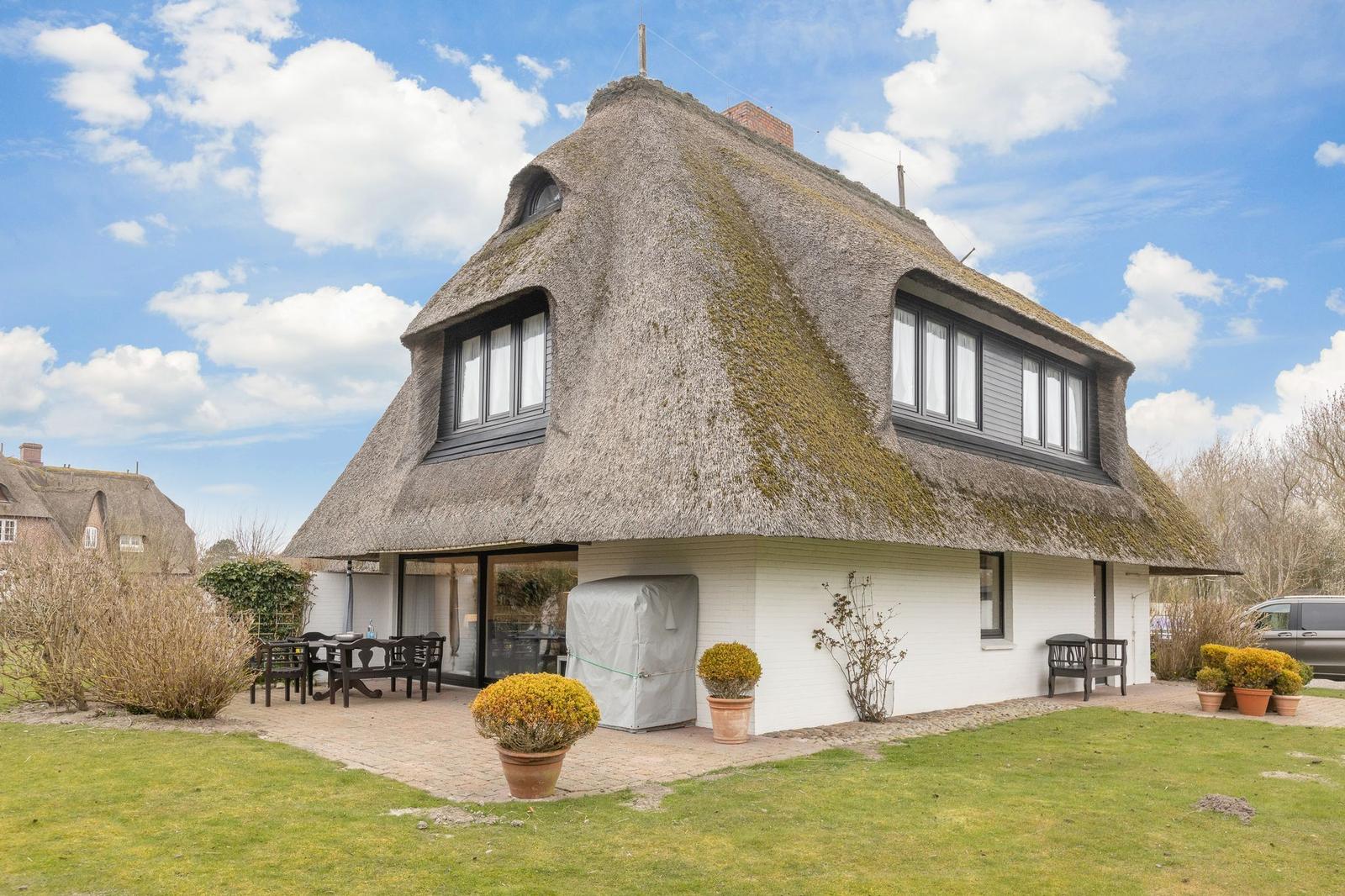 Haus mit strohgedecktem Dach und weißer Fassade. Außenbereich mit Tisch, Stühlen und Pflanzen.