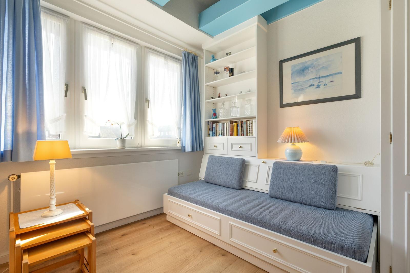 Zimmer mit Fenster, Bücherregal und Sitzbank mit blauen Kissen.