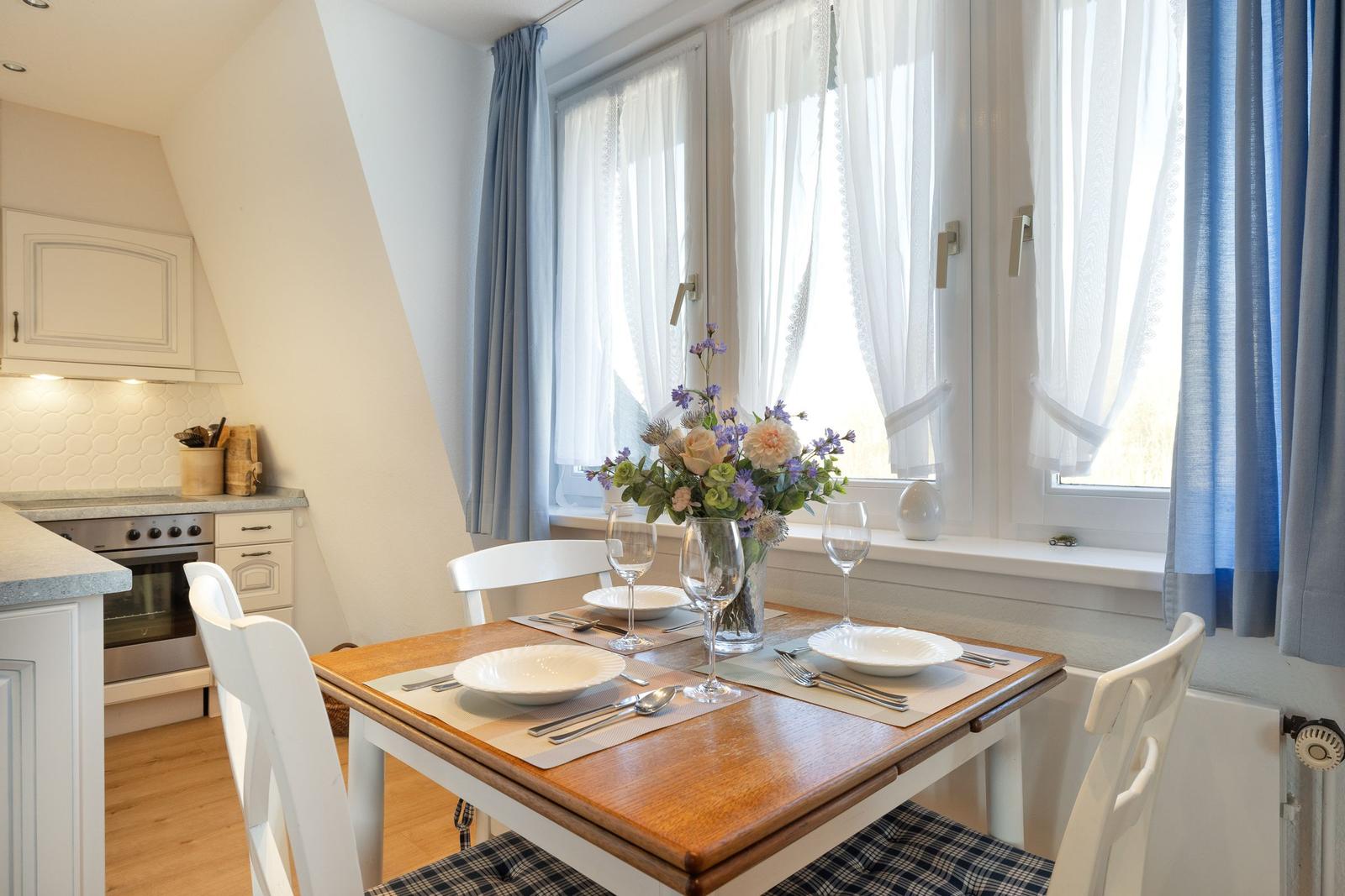 Essbereich mit Tisch, Stühlen und Blumen. Fenster mit weißen und blauen Vorhängen.