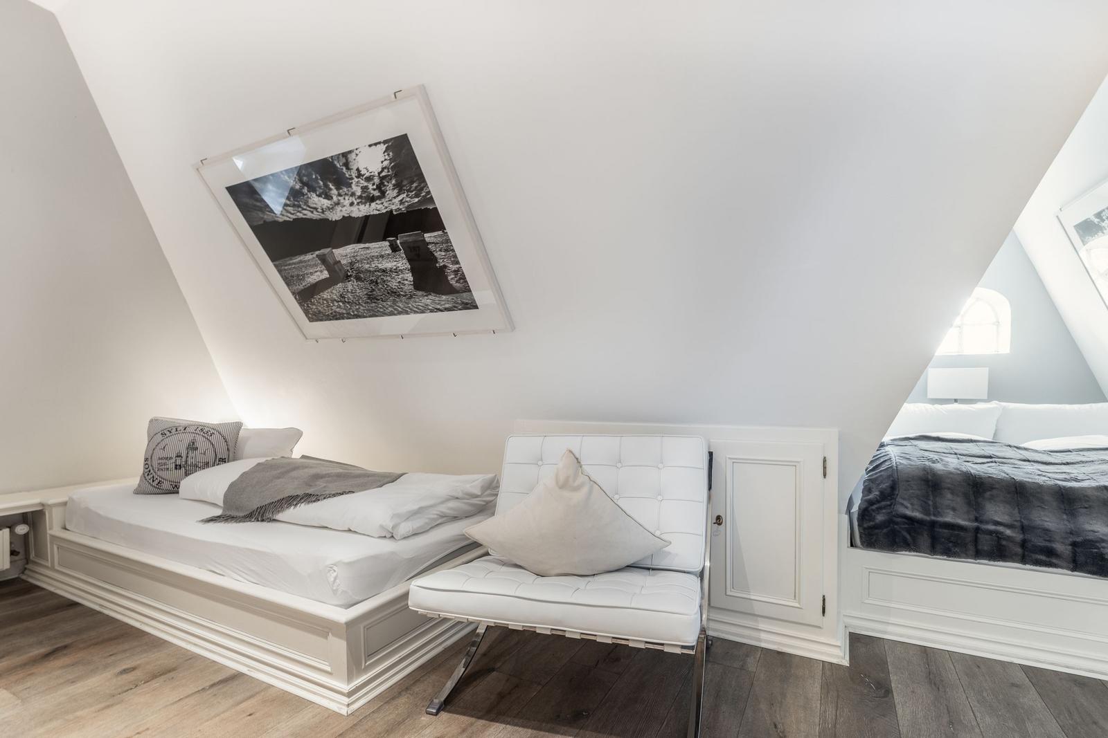 Room with two beds, white chair, and wall art under sloped ceiling.