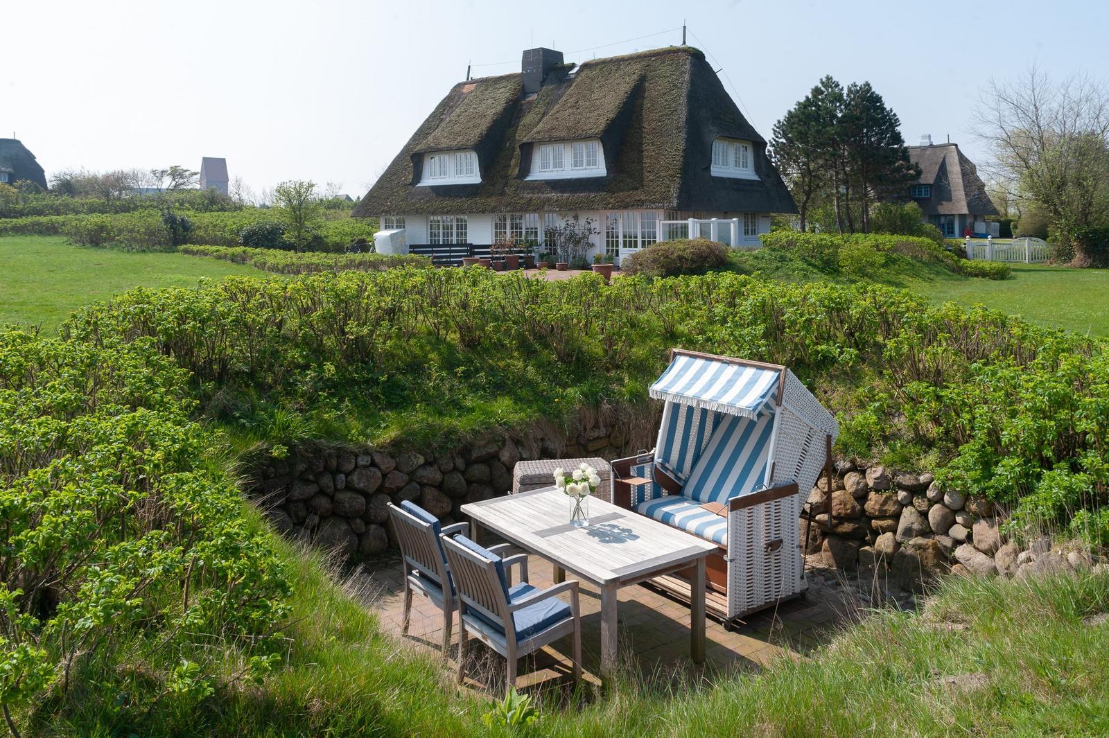 Terrasse mit Strandkorb und Tisch. Hintergrund: traditionelle Strohdachhäuser.