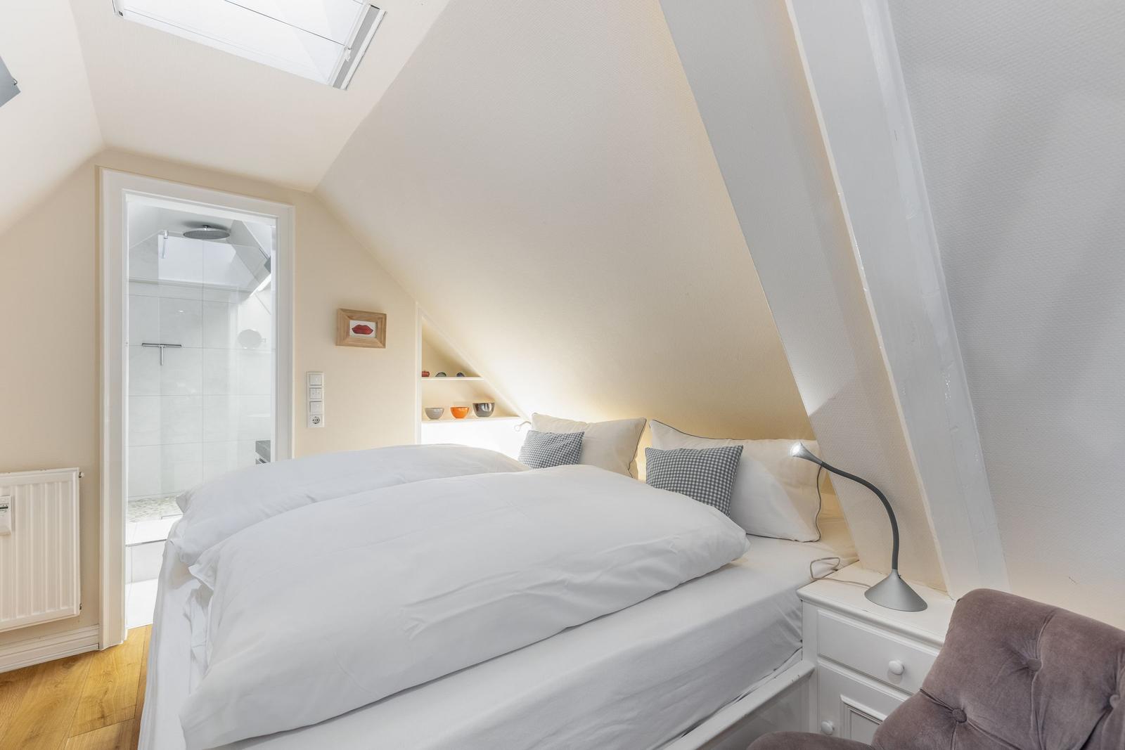 Schlafzimmer mit Dachschrägen, Bett, Nachttisch und Sessel. Durchgang zur Dusche.